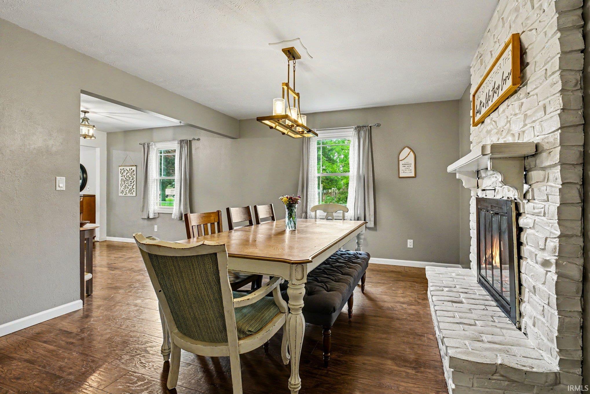 Dining area featuring a whitewashed brick fireplace with a mantel, wood-finish flooring, and a contemporary chandelier