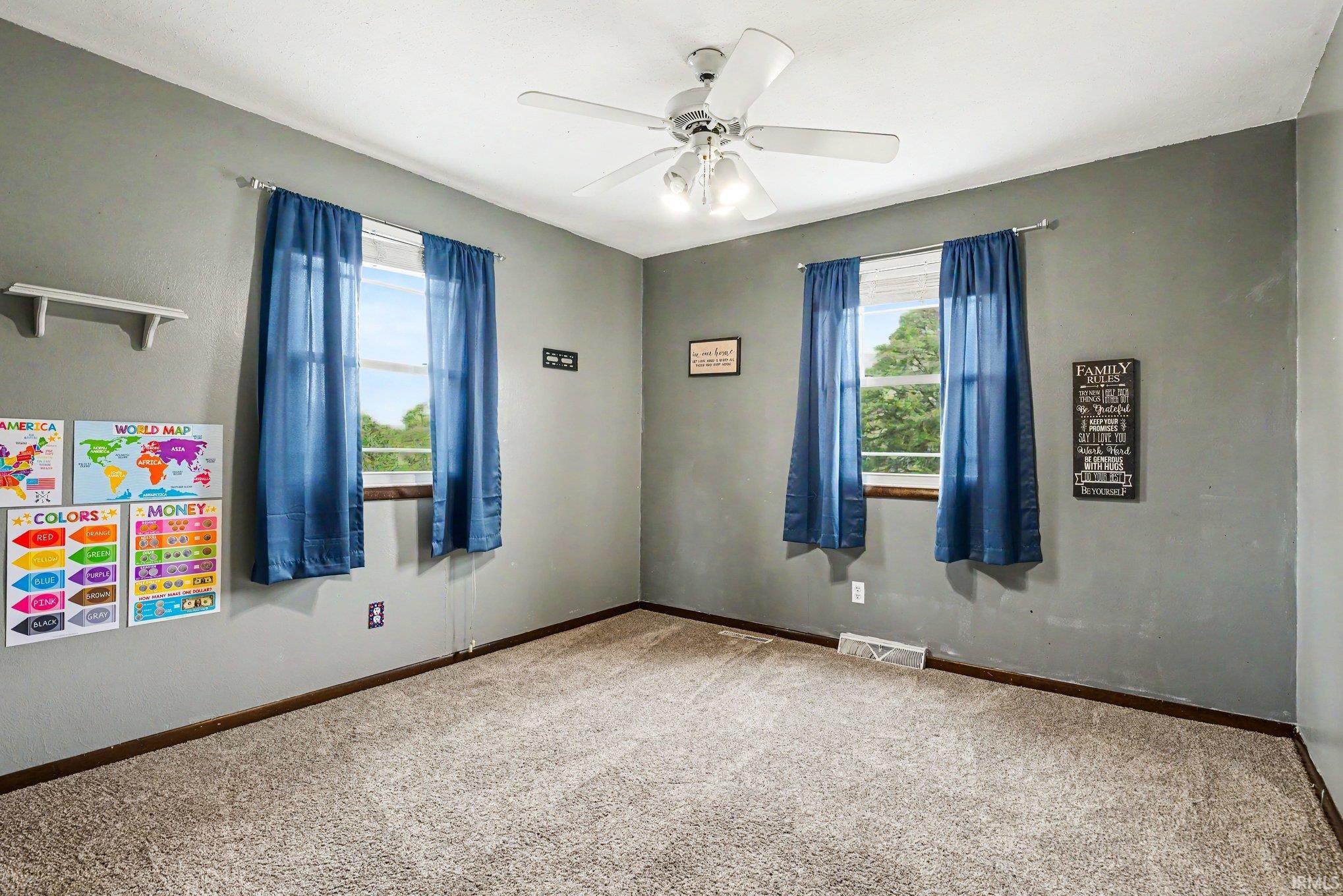 Carpeted room featuring gray walls, two windows with blue drapes, a white ceiling fan with integrated lighting, and white baseboards