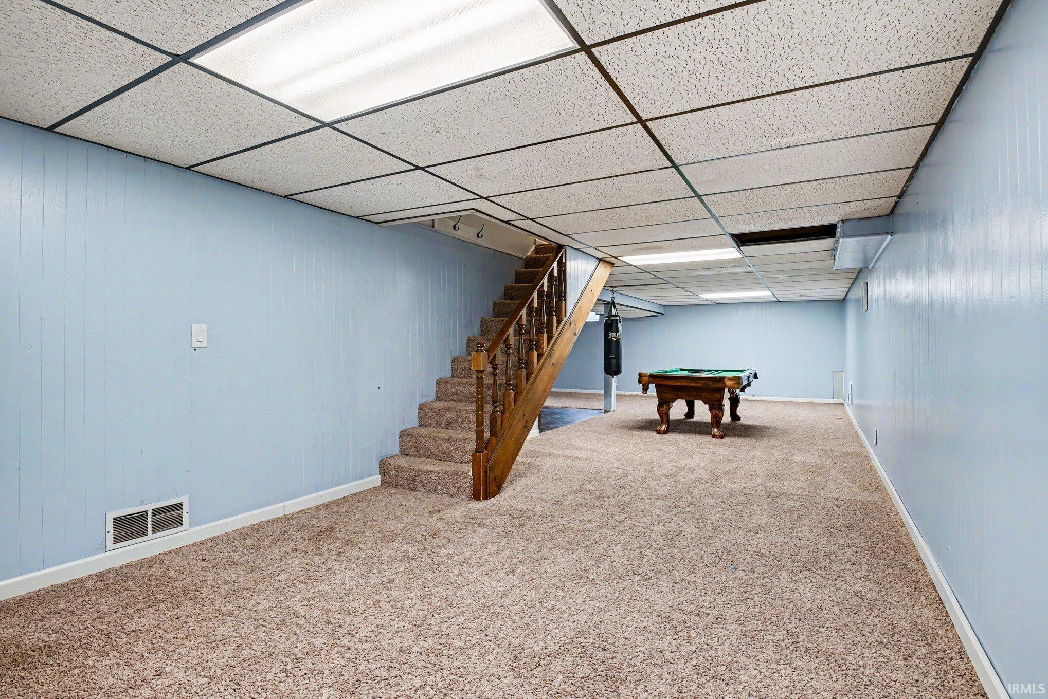 Spacious finished basement featuring light blue paneling walls, neutral carpet flooring, and a drop ceiling with integrated lighting