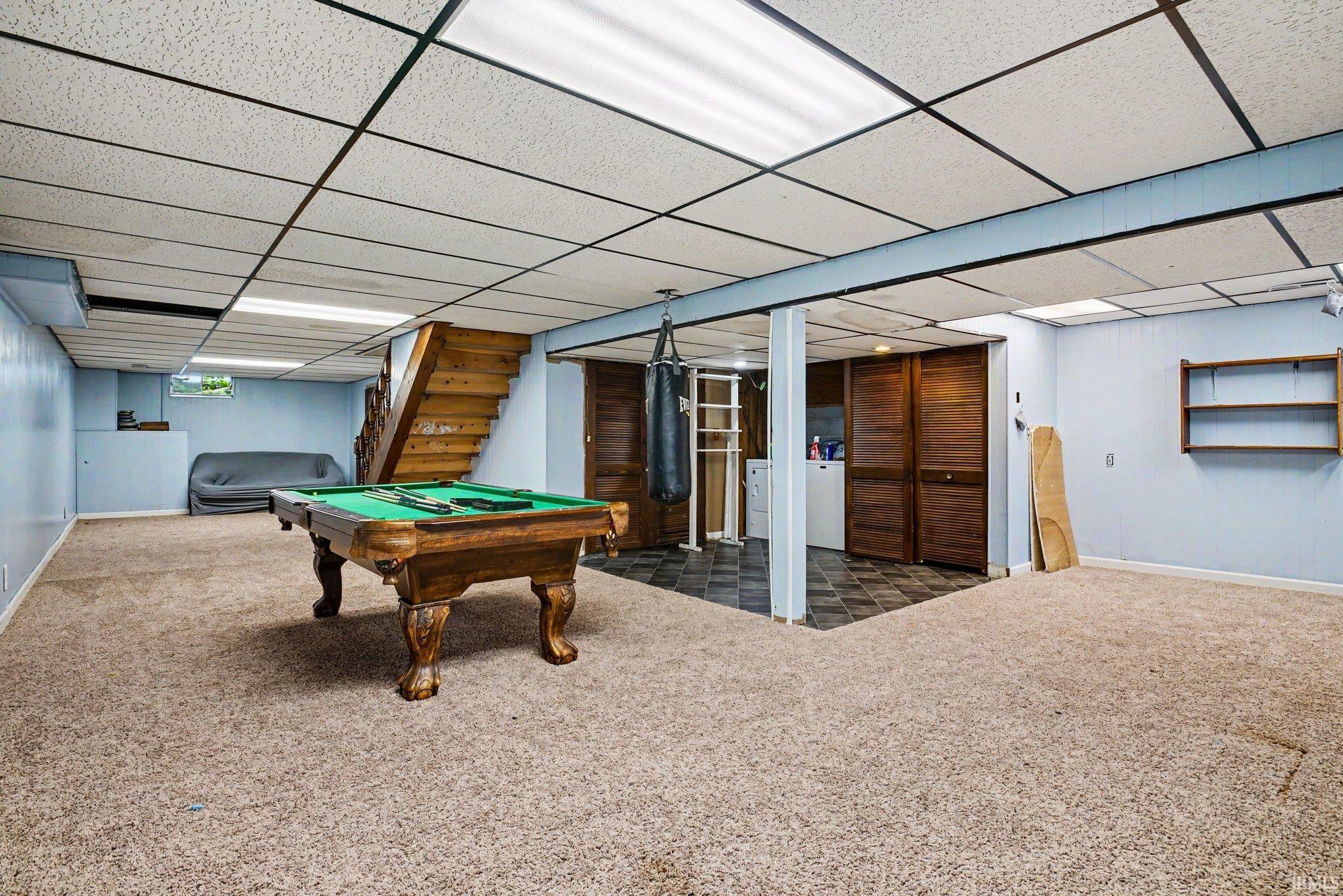 Carpeted lower level featuring a drop ceiling with integrated lighting, exposed wooden staircase, and built-in storage with bi-fold doors