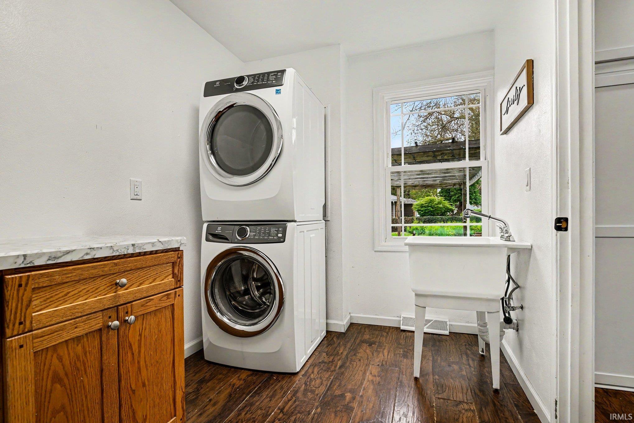 Dedicated laundry space featuring a stackable washer and dryer, wood-finish flooring, a utility sink, and built-in cabinetry with a stone-finish countertop