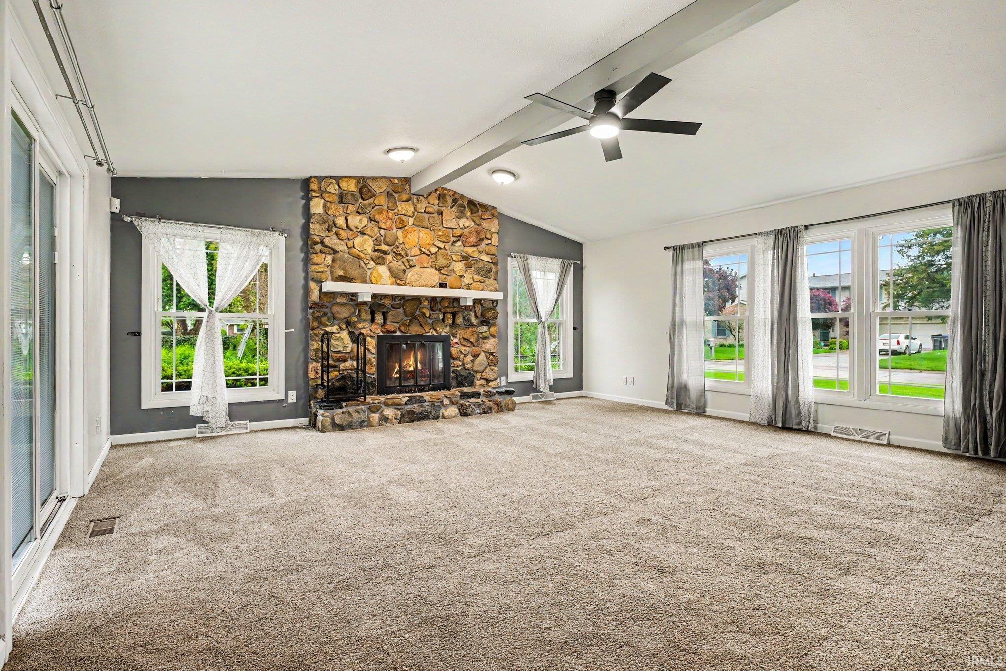Spacious living area featuring a stone-clad fireplace with a white mantel, vaulted ceilings with exposed beam, and a ceiling fan