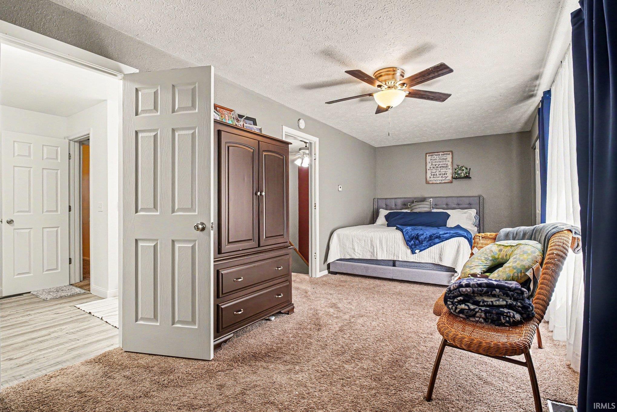 Carpeted room with a ceiling fan with integrated lighting, window with blue and white drapes, and an adjacent hallway with wood-finish flooring