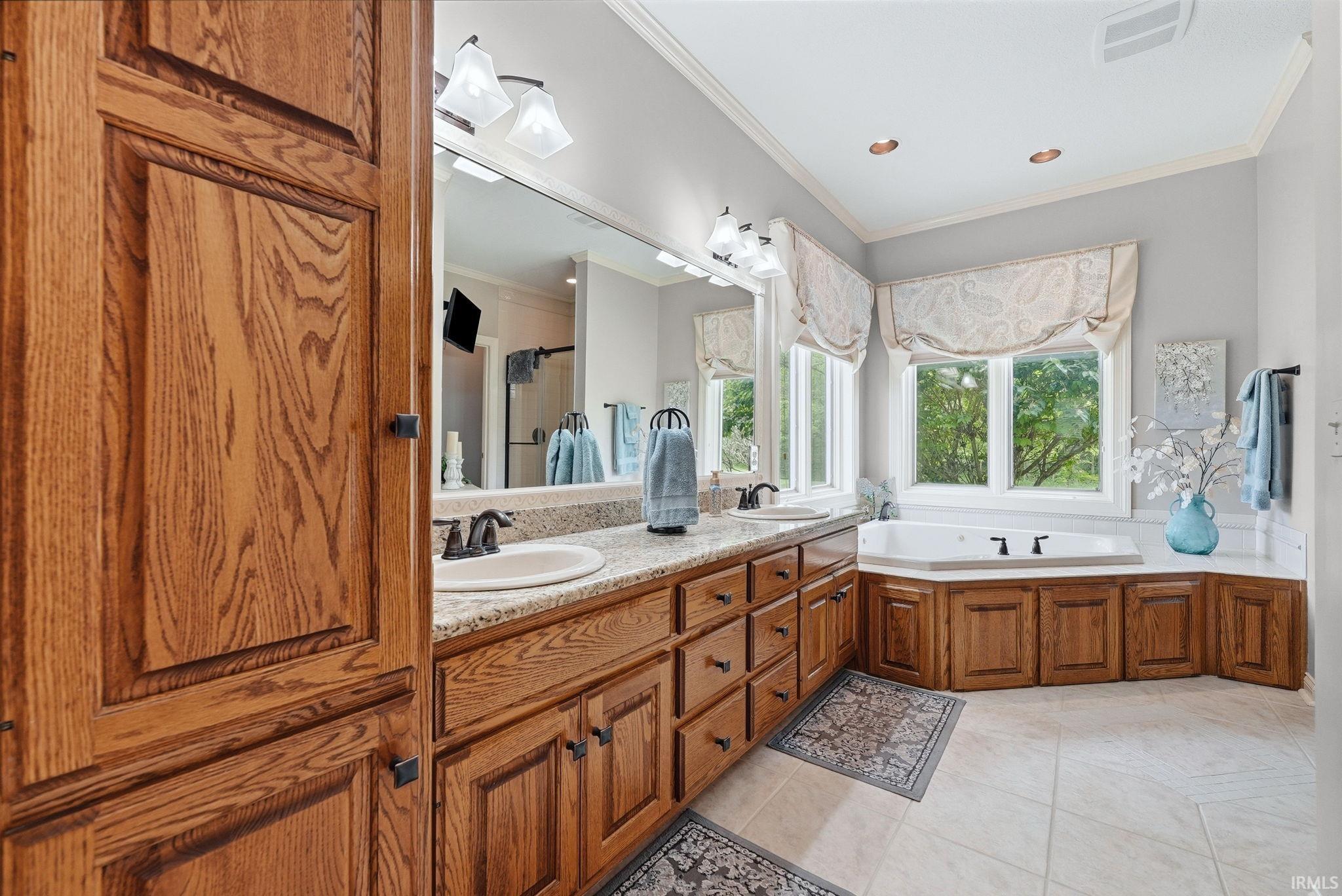 Spacious bathroom featuring a dual vanity with granite countertops, rich wood cabinetry offering ample storage, an integrated corner soaking tub, and multiple windows providing natural light