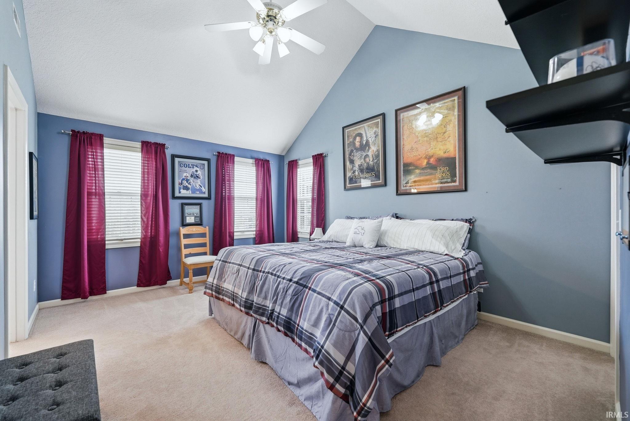 Room featuring a vaulted ceiling with a ceiling fan, light beige carpet flooring, multiple windows with blinds, and built-in wall shelving