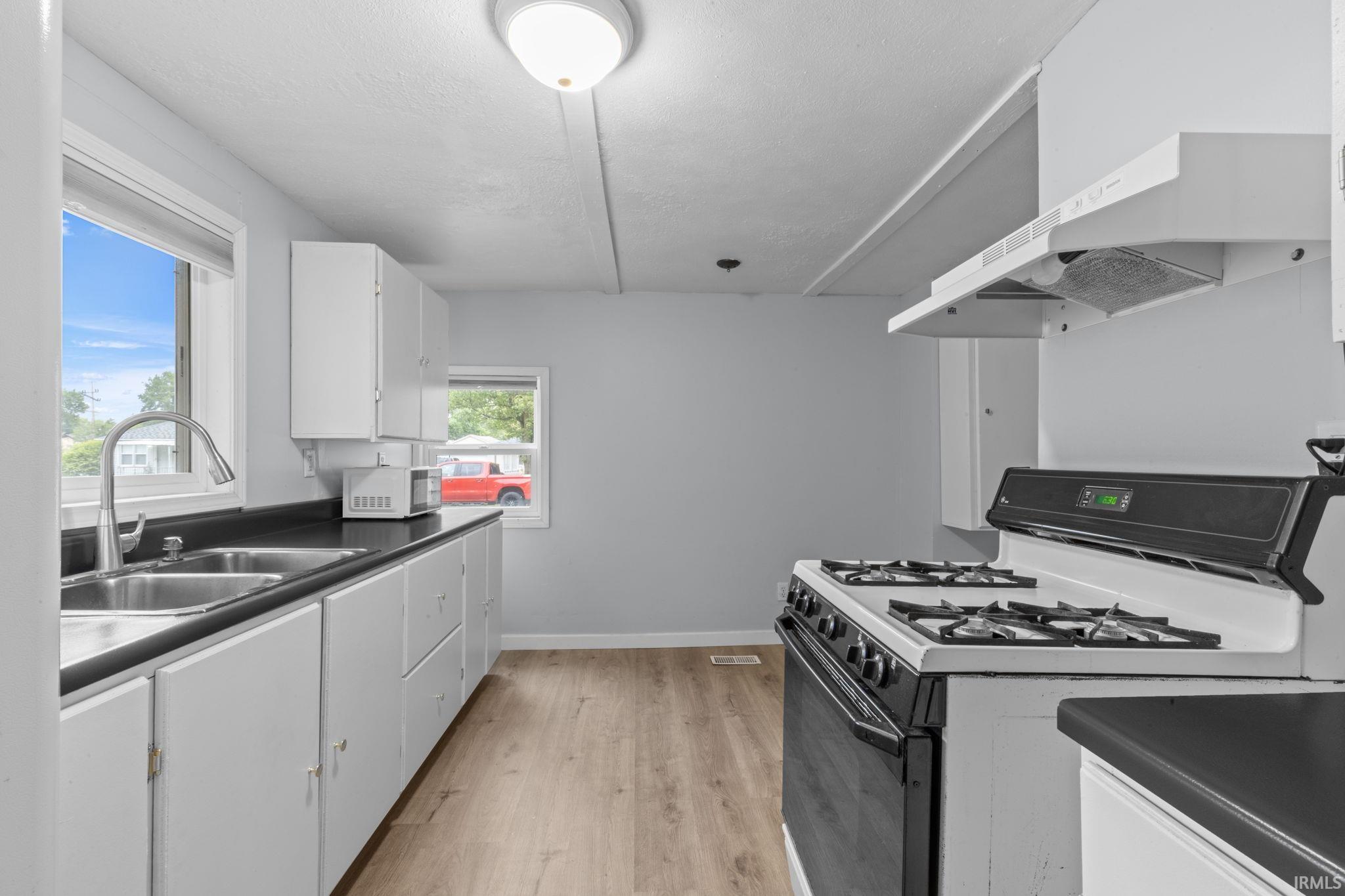 Kitchen featuring wood-finish flooring, white cabinetry, a double basin stainless steel sink with a gooseneck faucet, black countertops, and a white and black range with an overhead exhaust hood