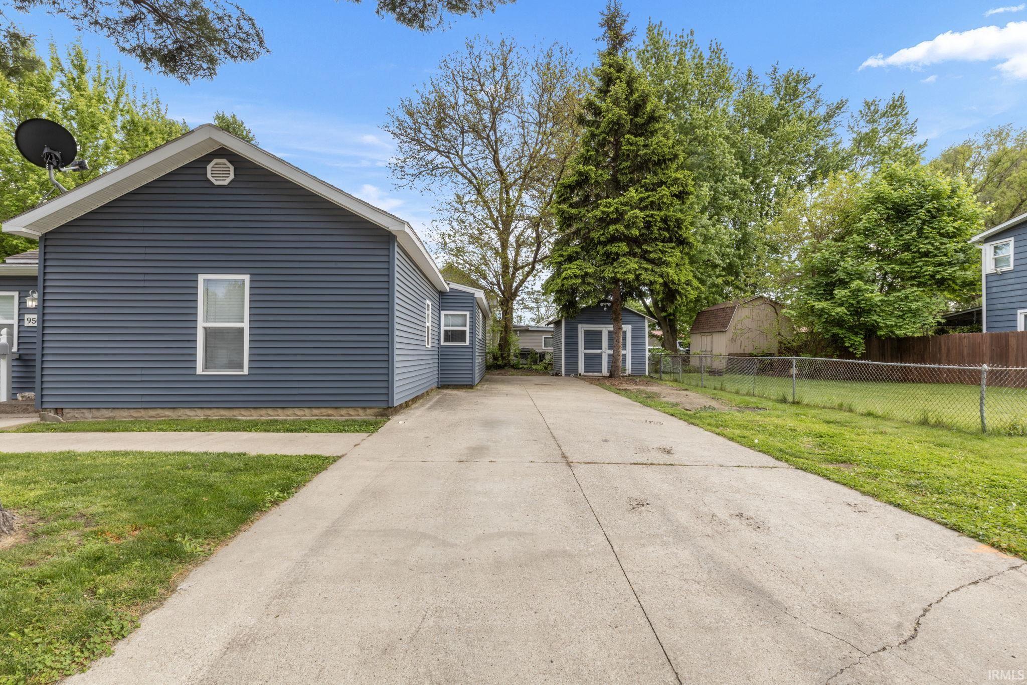 Concrete driveway leading to a blue siding residence featuring white trim, a side entry, and a backyard with a chain-link fence and a small shed