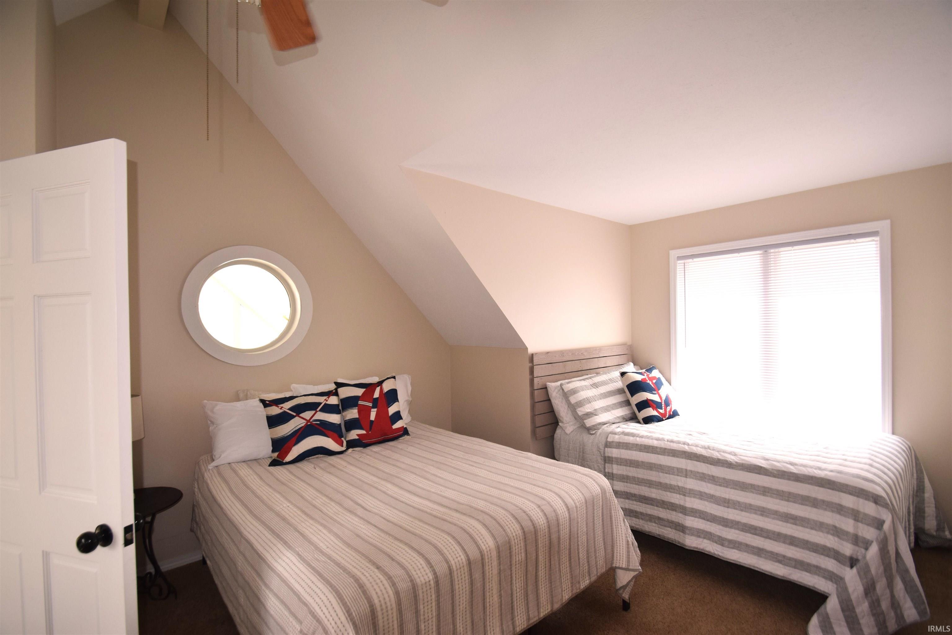 Sloped ceiling room featuring a circular window, rectangular window with blinds, panel door with dark hardware, and a ceiling fan