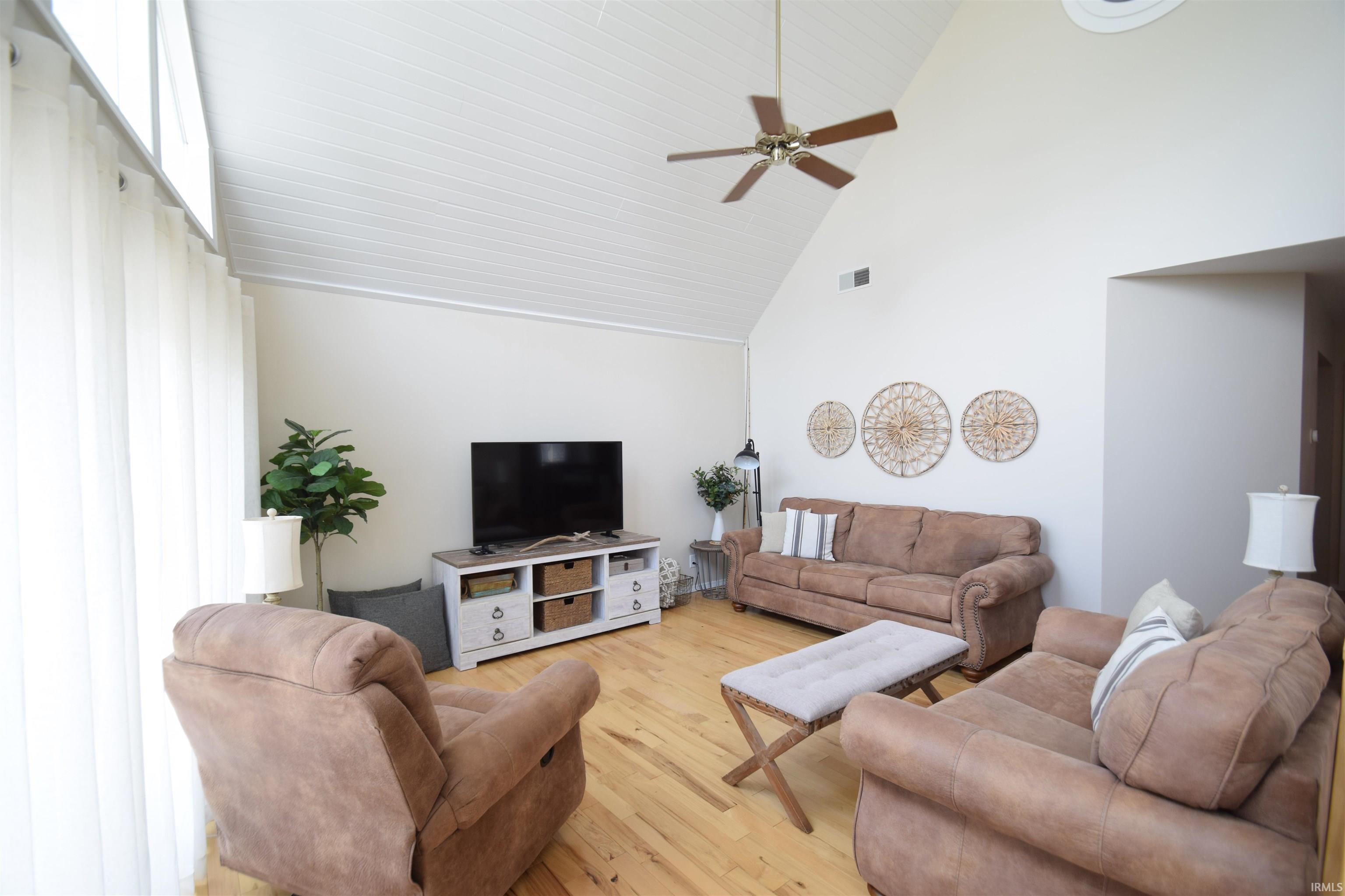 Spacious living area featuring a vaulted tongue-and-groove ceiling with a ceiling fan, abundant natural light from large windows, light wood-finish flooring, and a neutral color palette