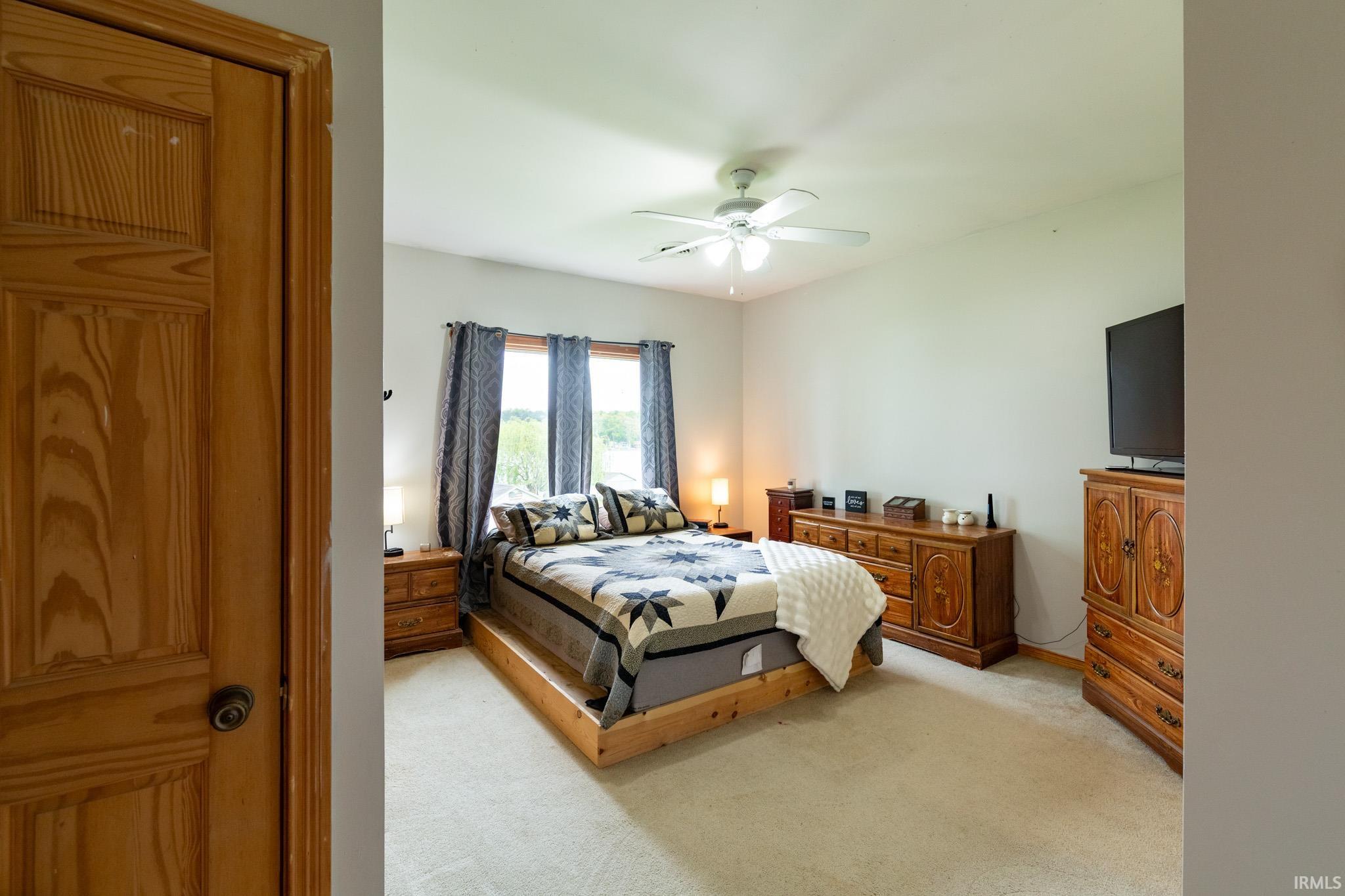 Carpeted bedroom featuring a ceiling fan with light fixture, three large windows, light-toned walls, and a natural wood-finish door with a dark knob