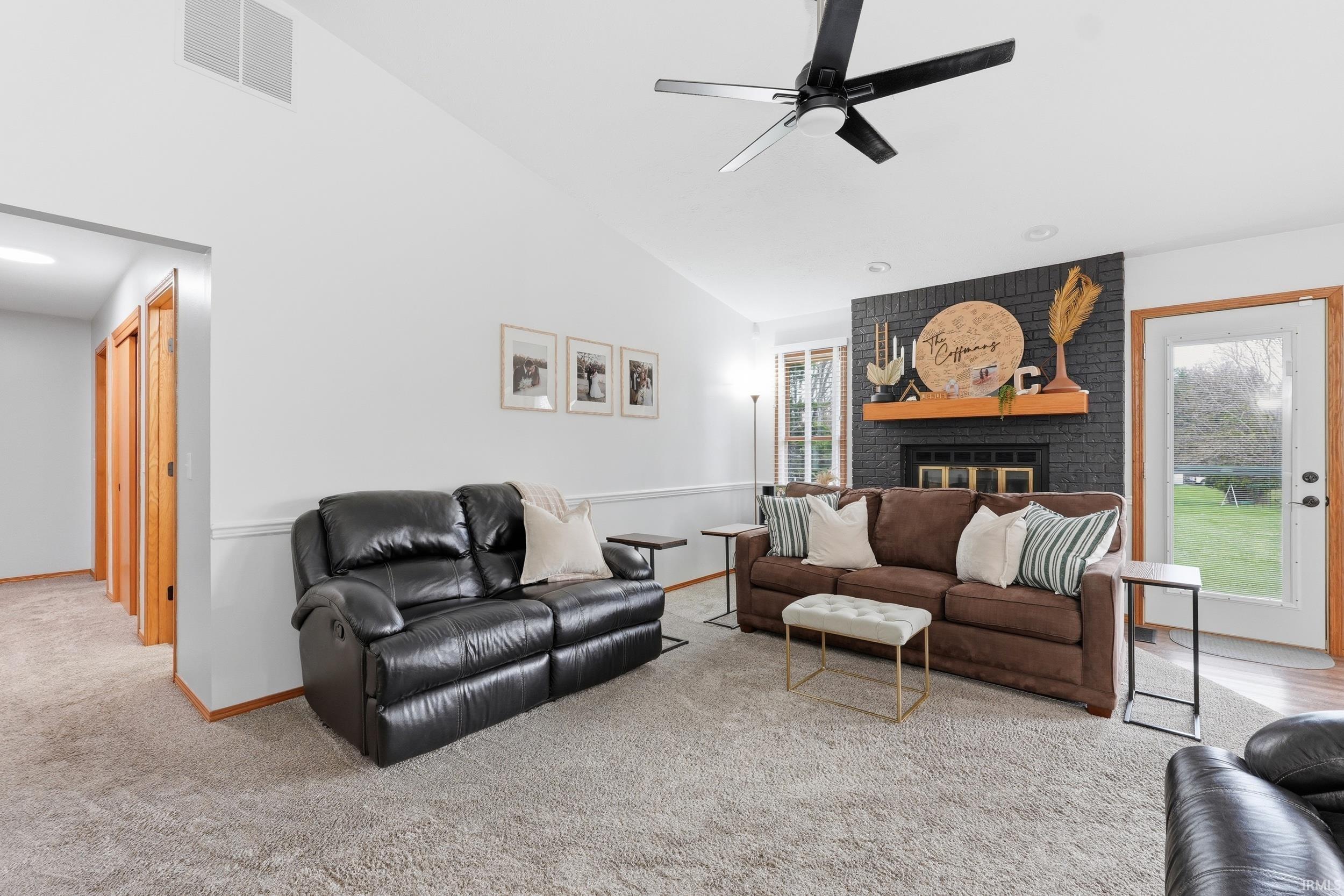 Carpeted living area featuring vaulted ceiling, a brick fireplace, a ceiling fan, and healthy amount of natural light