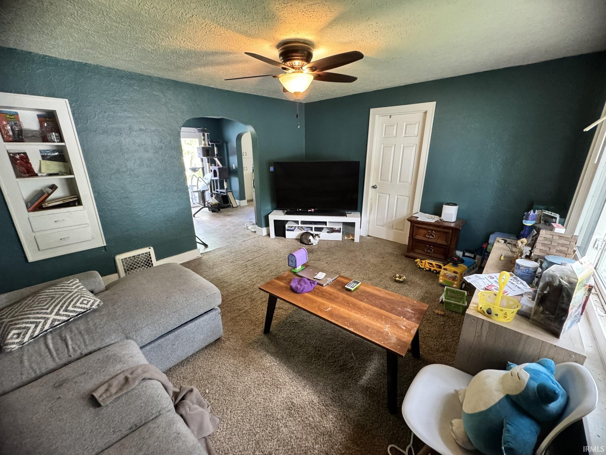 Carpeted room featuring a ceiling fan with integrated lighting, an arched entryway, and a built-in wall niche with shelving and lower cabinetry
