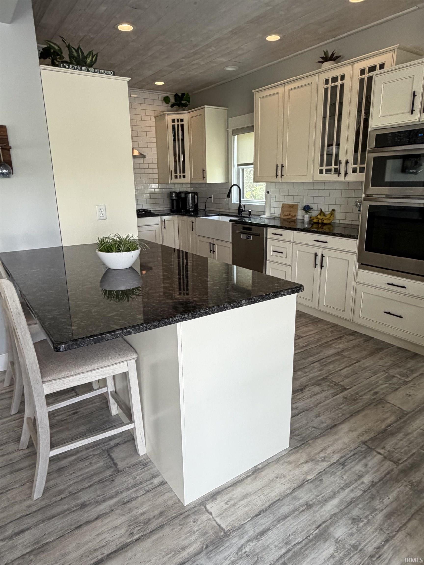 Kitchen featuring a central island with a dark granite countertop, light wood-finish flooring, white subway tile backsplash, and light-toned cabinetry with glass-front accents