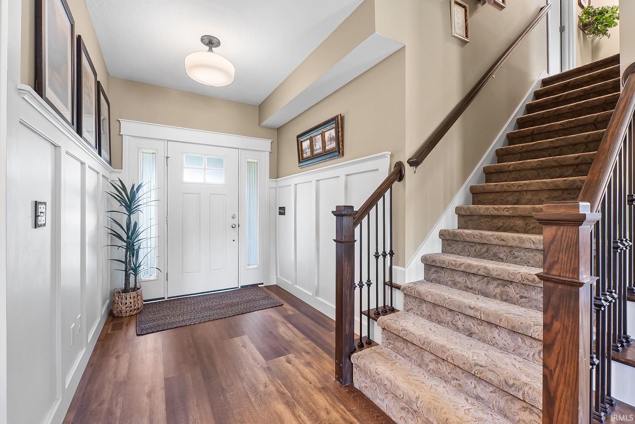 Entryway featuring a white paneled door with glass inserts, wood-finish flooring, white wainscoting, and a staircase with dark wood newel posts, iron balusters, and carpeted treads