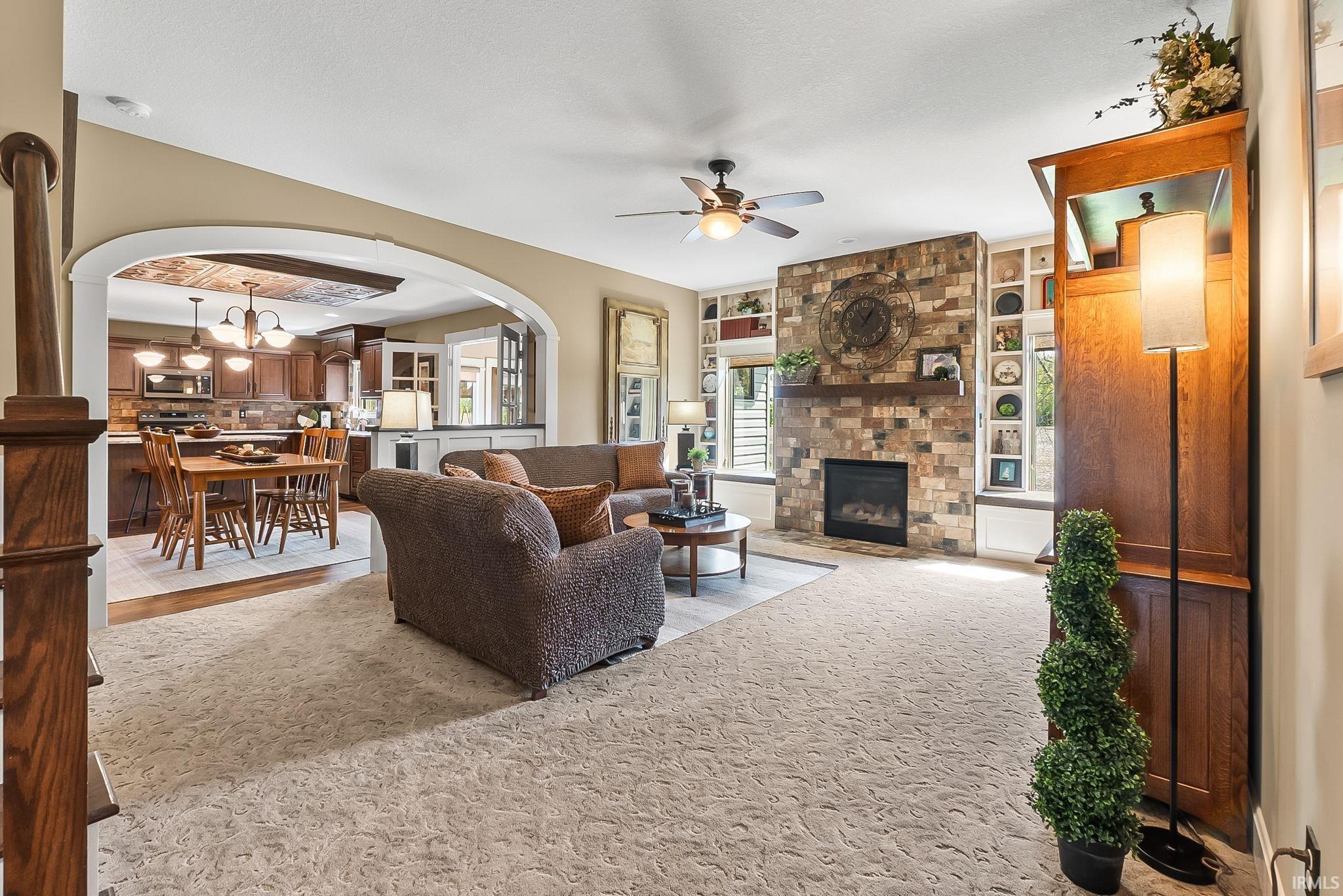 Living room featuring a stacked stone fireplace with wood mantel, built-in shelving, a ceiling fan, and patterned carpeting