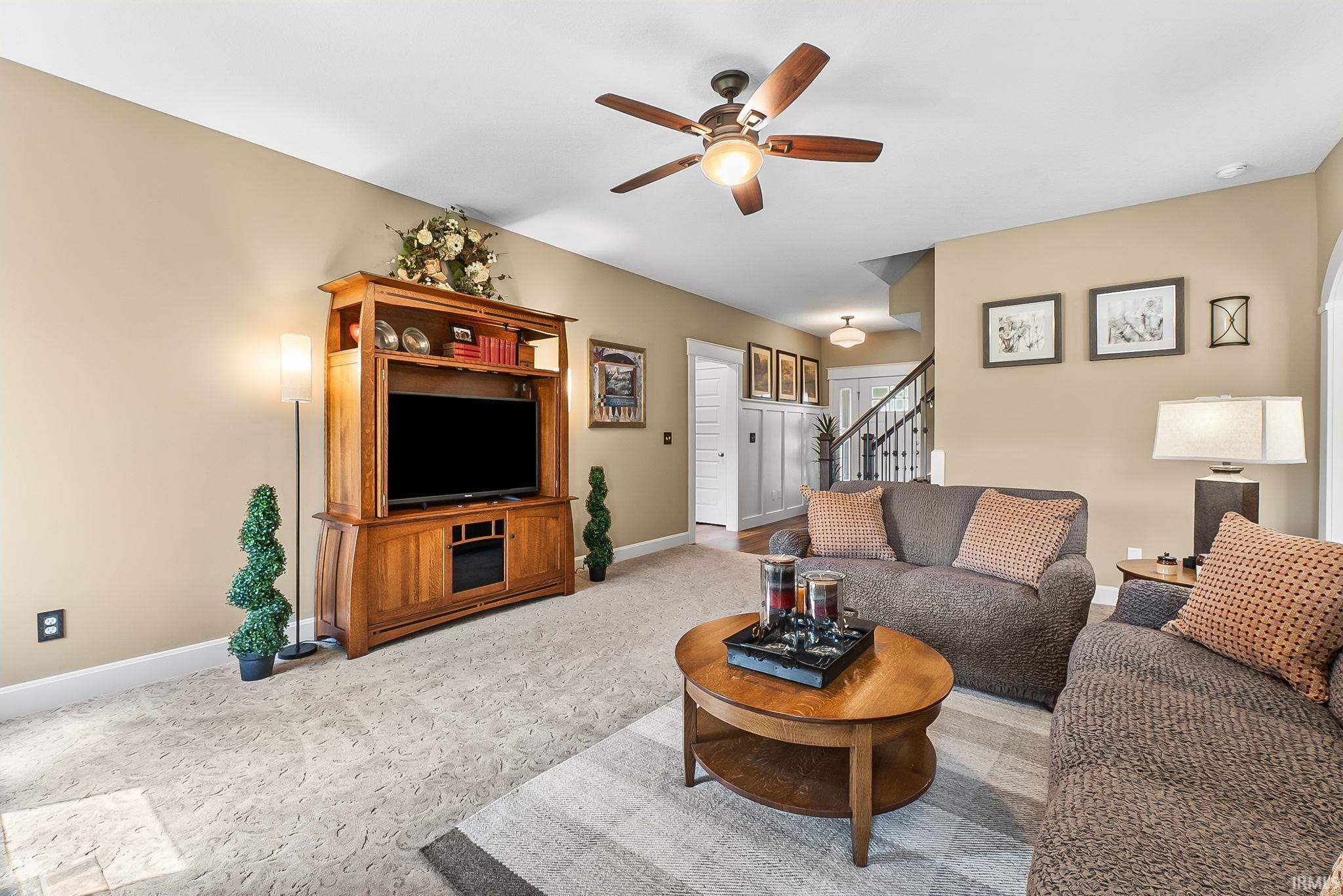 Living area featuring neutral wall tones, patterned carpeting, a ceiling fan, and a visible staircase with dark railings