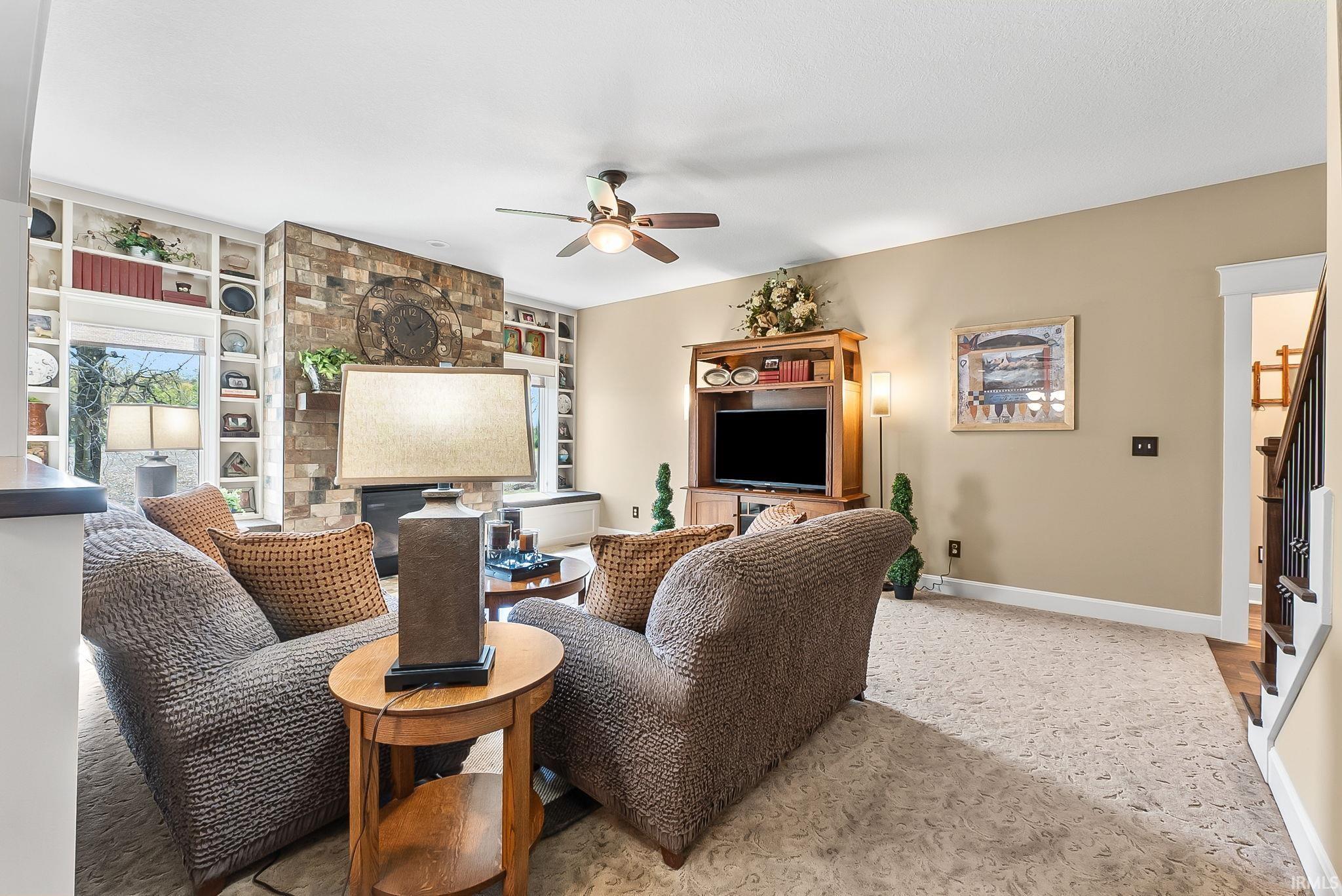 Living area showcasing a stone-clad fireplace, integrated shelving, large windows, ceiling fan, and hardwood flooring on the stairway