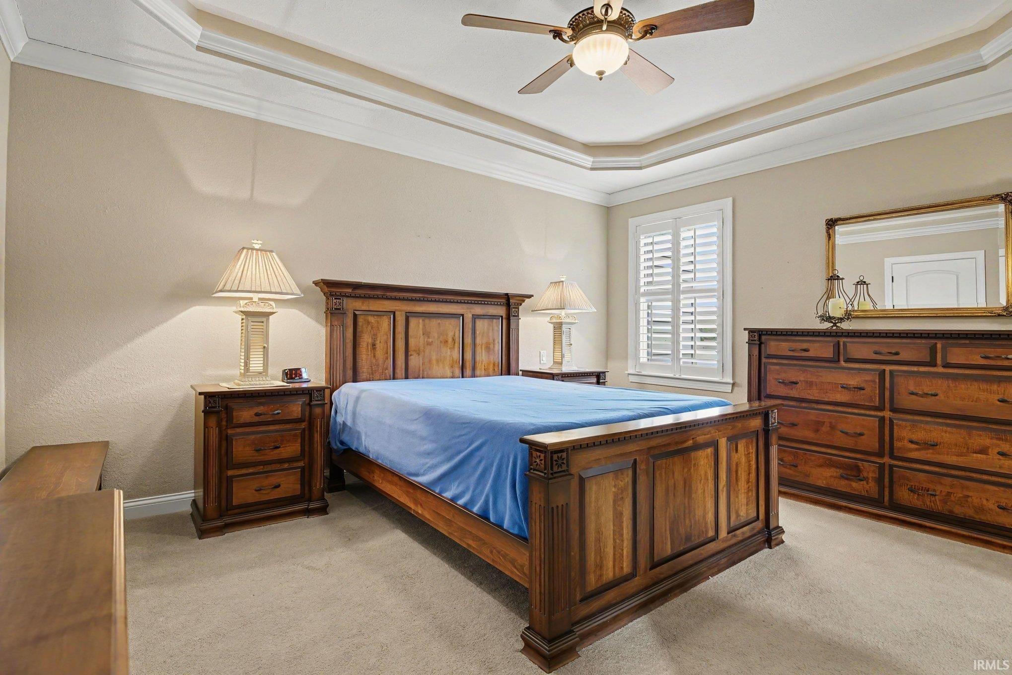 Recessed tray ceiling with crown molding, ceiling fan with integrated lighting, window with plantation shutters, and neutral carpet flooring