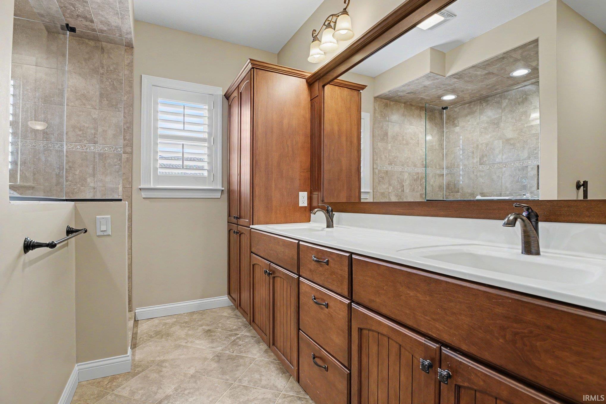 Bathroom featuring an expansive wood-finish vanity with dual sinks and white countertops, a large mirror, a tiled shower enclosure, and a window with plantation shutters