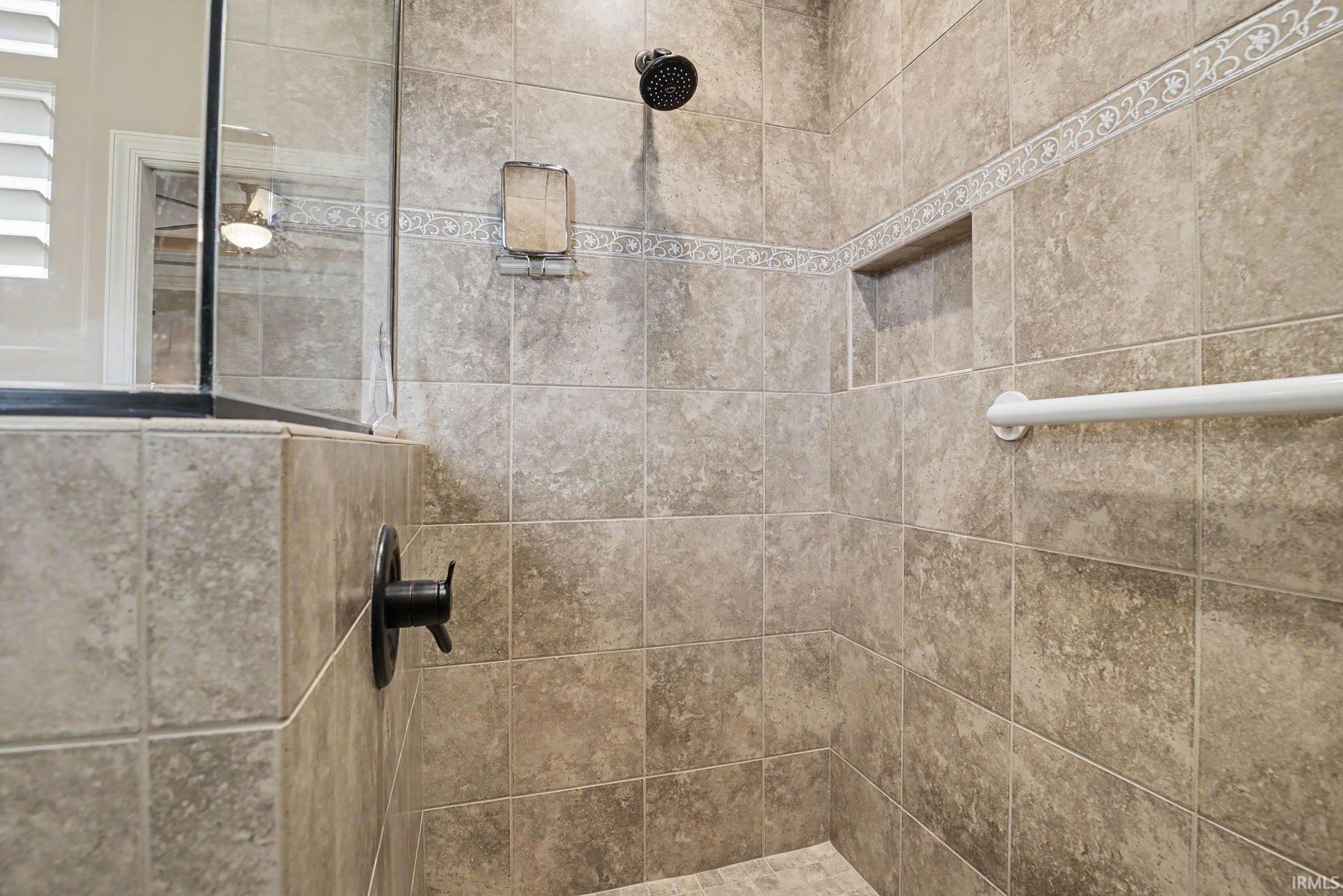 Shower enclosure featuring neutral-toned tile walls with a decorative accent border, a built-in shower niche, a dark-finished shower head, and a white grab bar