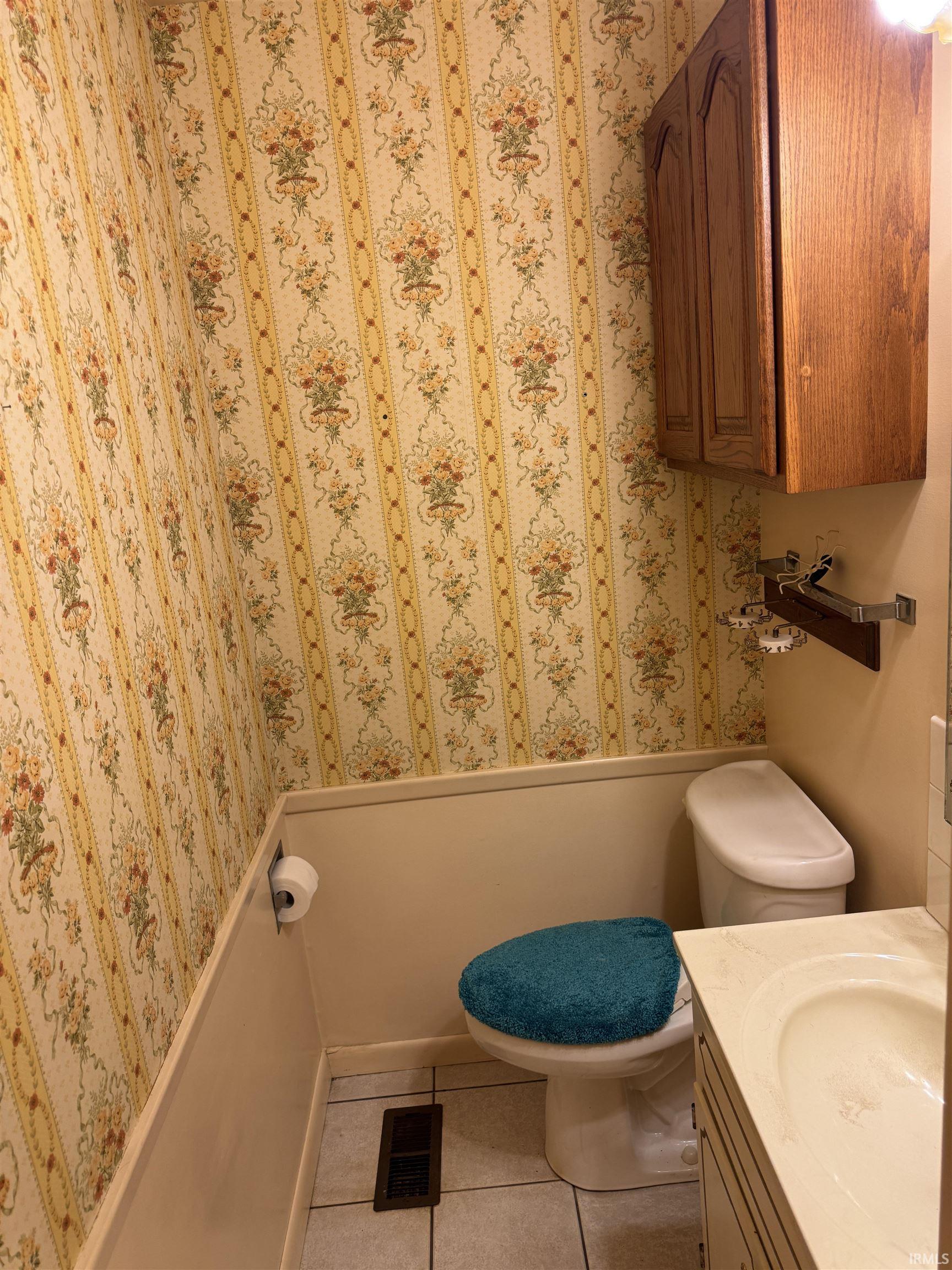 Compact bathroom featuring floral wallpaper, wainscoting, a white toilet, a vanity with an integrated sink, and wood-finish cabinetry