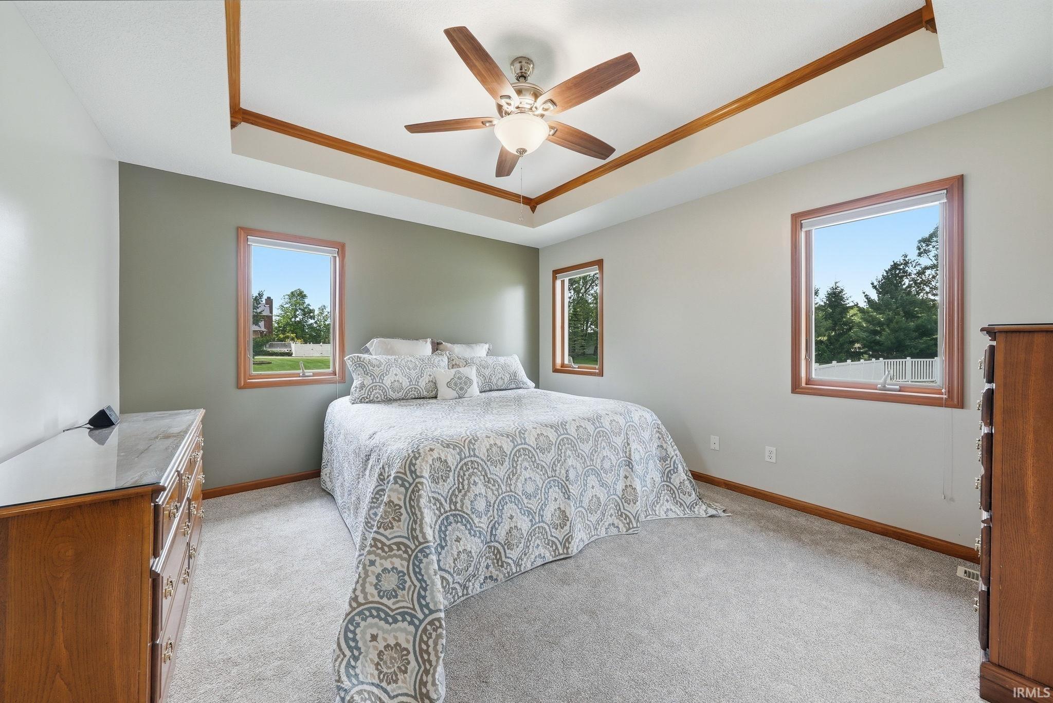 Bedroom featuring a tray ceiling with wood trim, a ceiling fan, light gray carpeting, and multiple windows with wood-framed casements