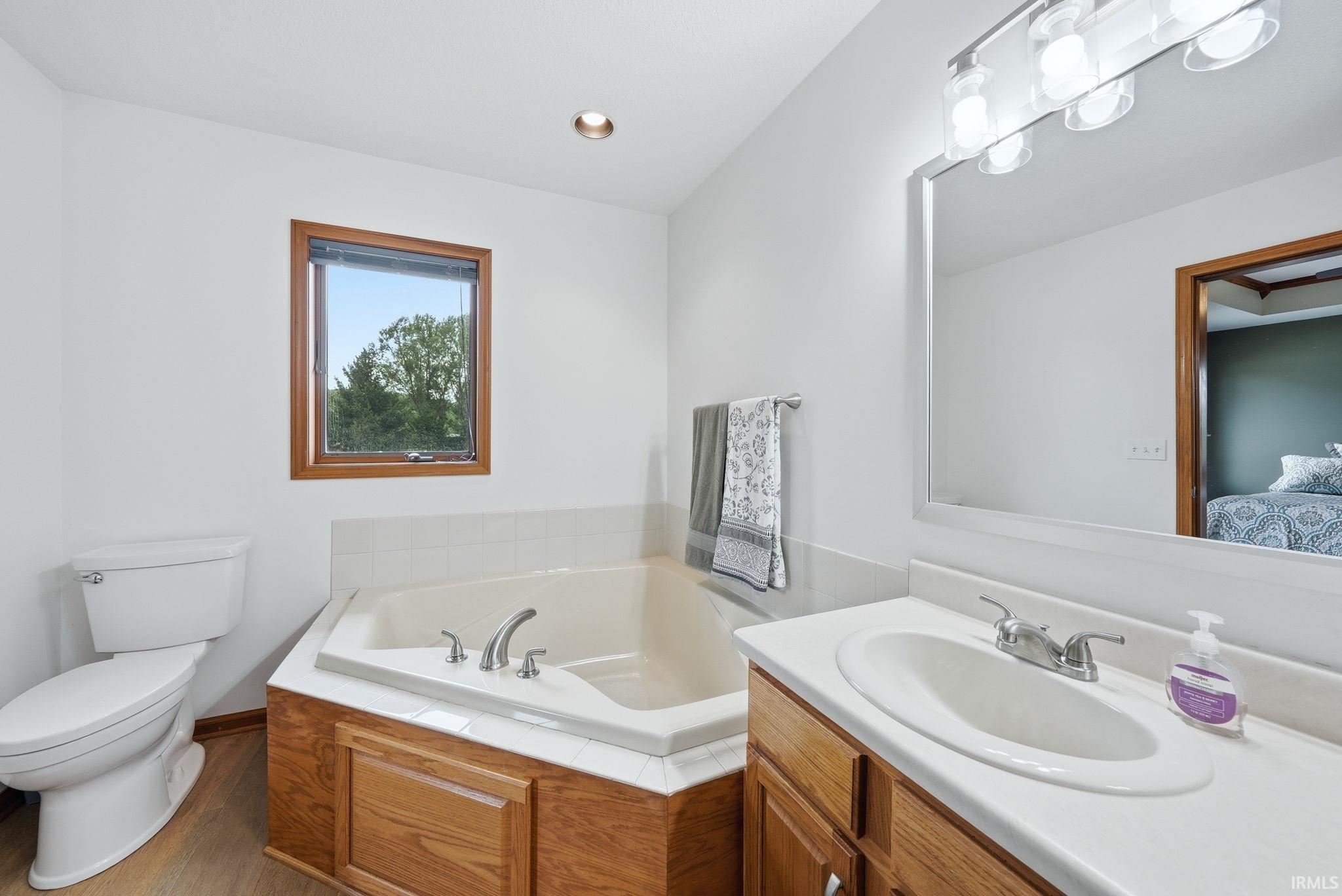 Bathroom featuring a corner built-in tub with tile surround, a single vanity with an oval basin, and wood-finish flooring