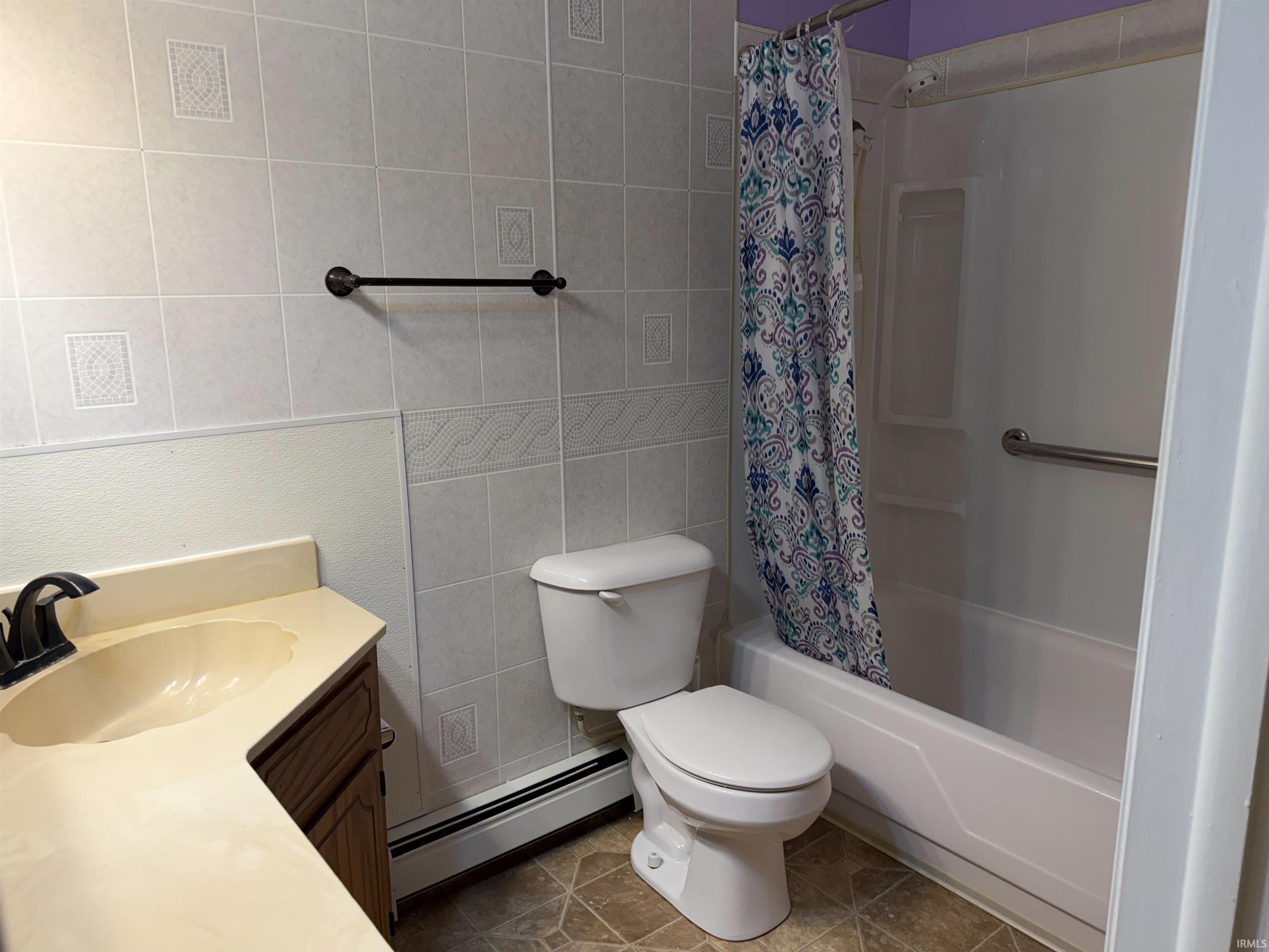 Bathroom with light-toned tiled walls, a built-in bathtub with shower, a vanity featuring a single-basin sink, and wood-finish flooring