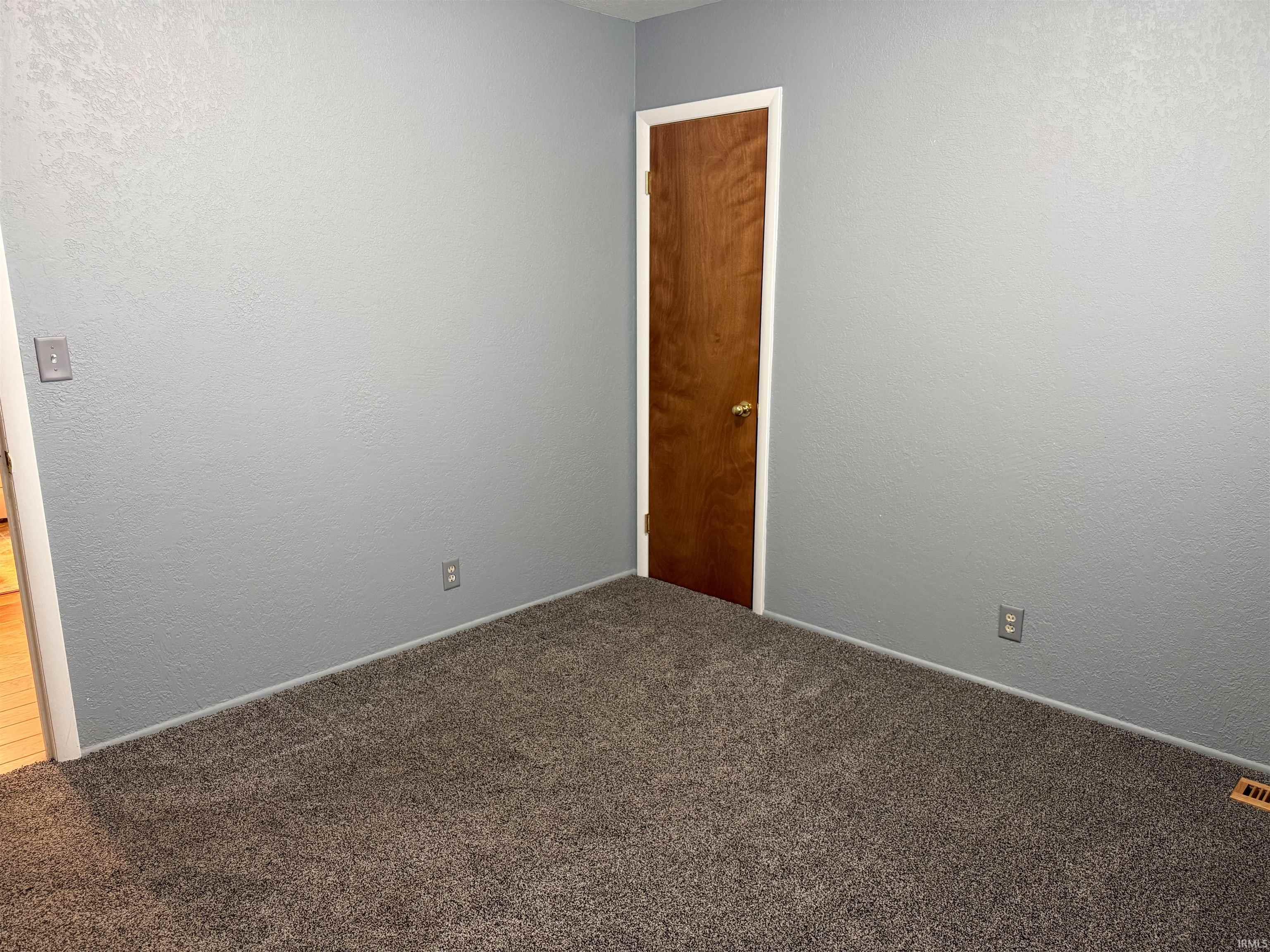Carpeted room featuring light gray walls, a wood-finish door with white trim, and a floor-level air vent