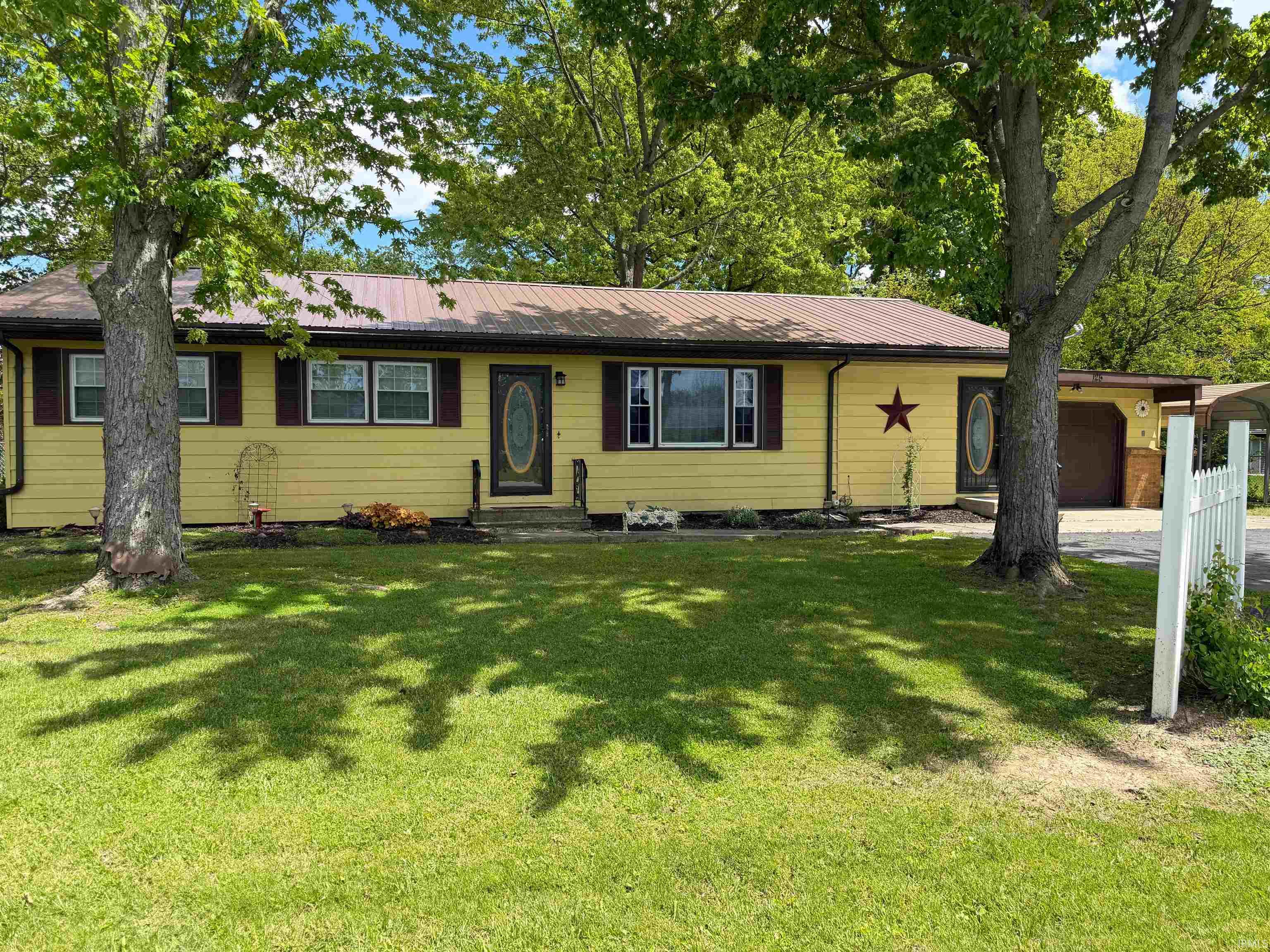 Yellow siding residence featuring a metal roof, dark brown shutters, and mature front yard trees