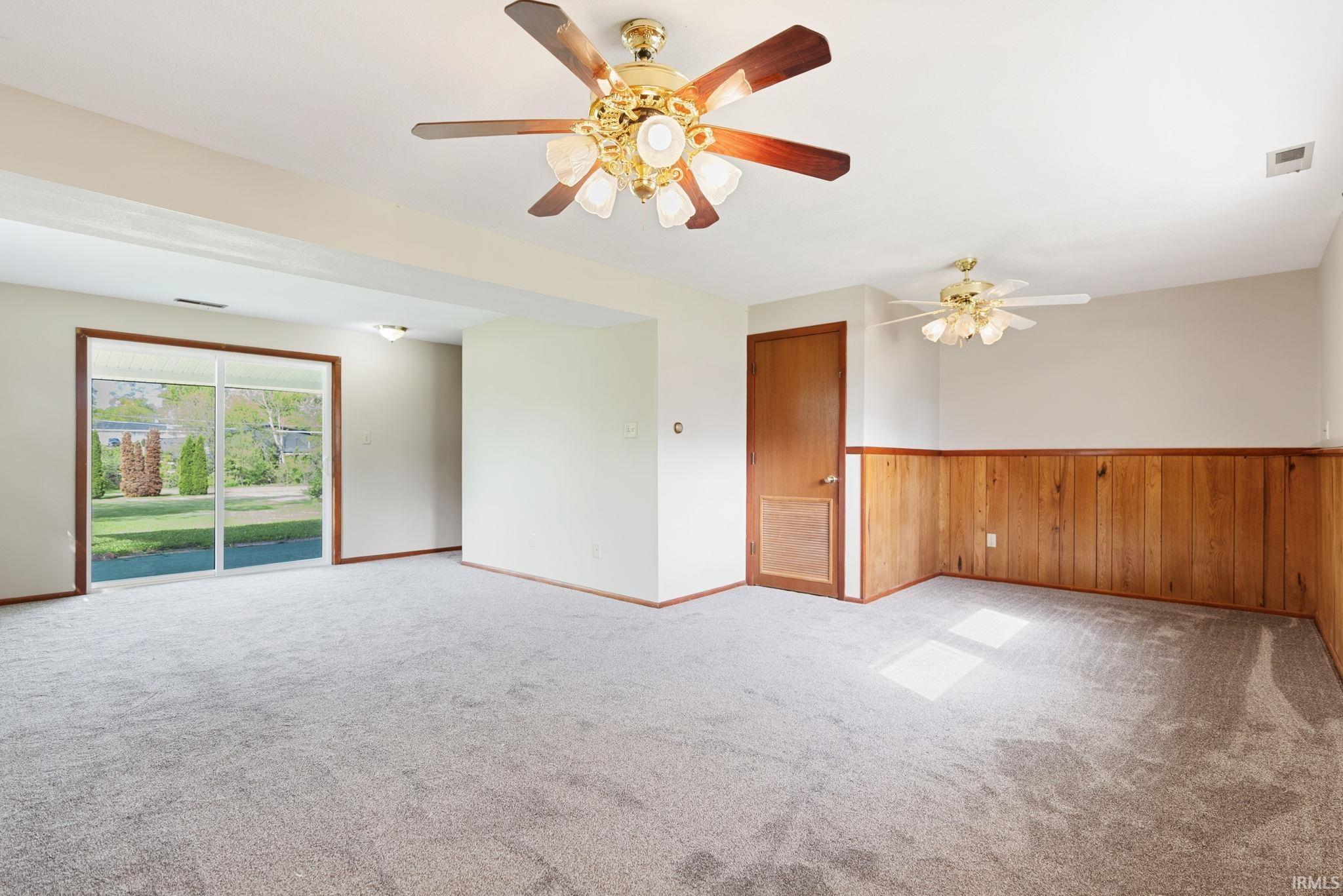 Spacious room featuring light gray carpeting, wood paneling, and a large sliding glass door opening to outdoor green space