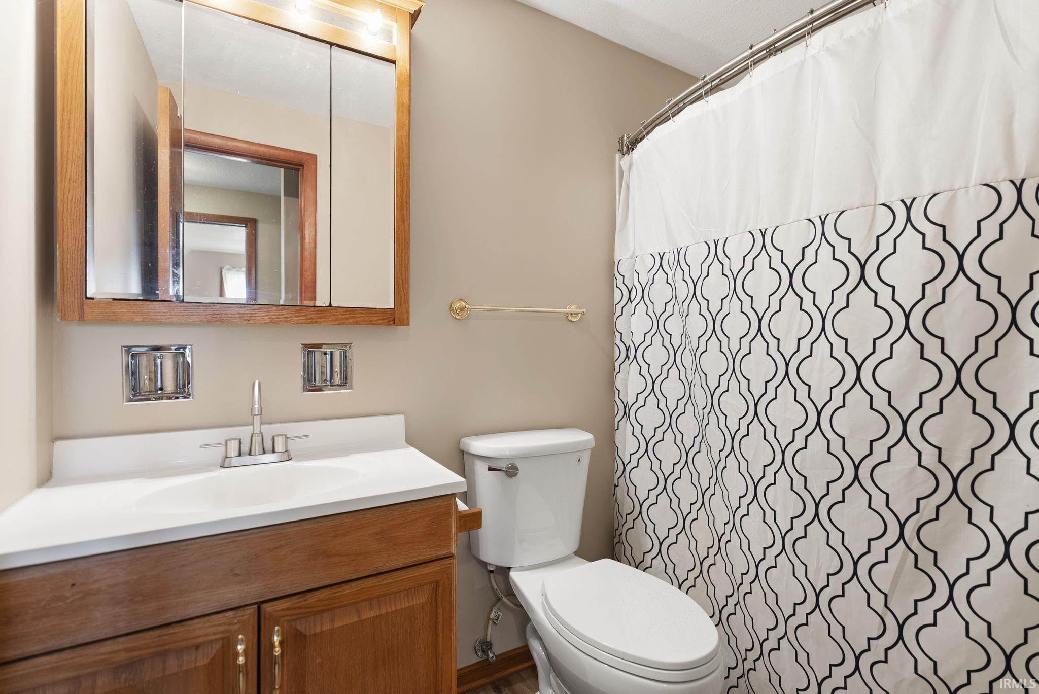 Bathroom featuring a wood-finish vanity with an integrated sink and white countertop, mirrored medicine cabinet, toilet, and shower with a patterned curtain