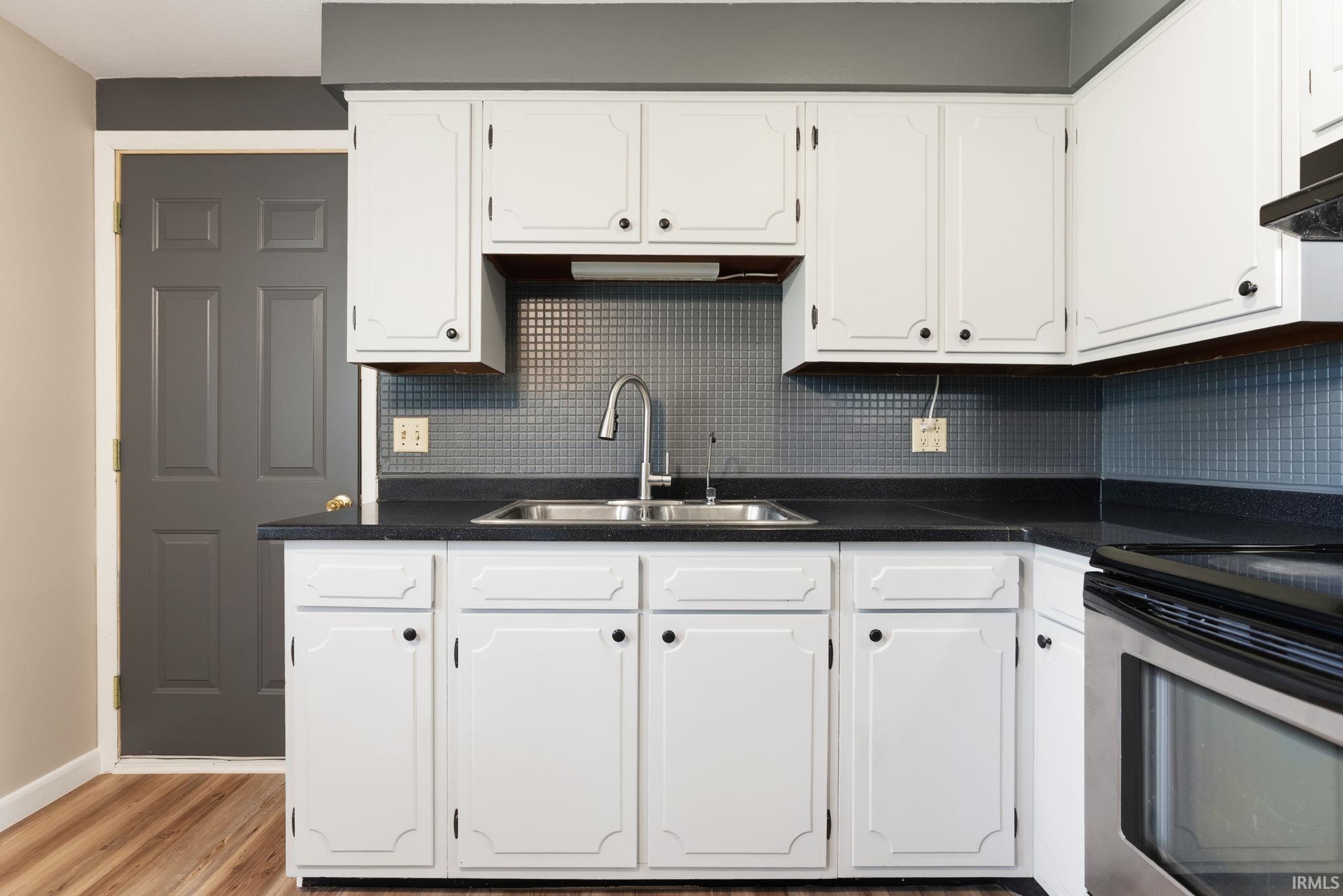 Kitchen featuring white cabinetry with black hardware, dark countertops, a stainless steel double-basin sink with a gooseneck faucet, a tile backsplash, and wood-finish flooring