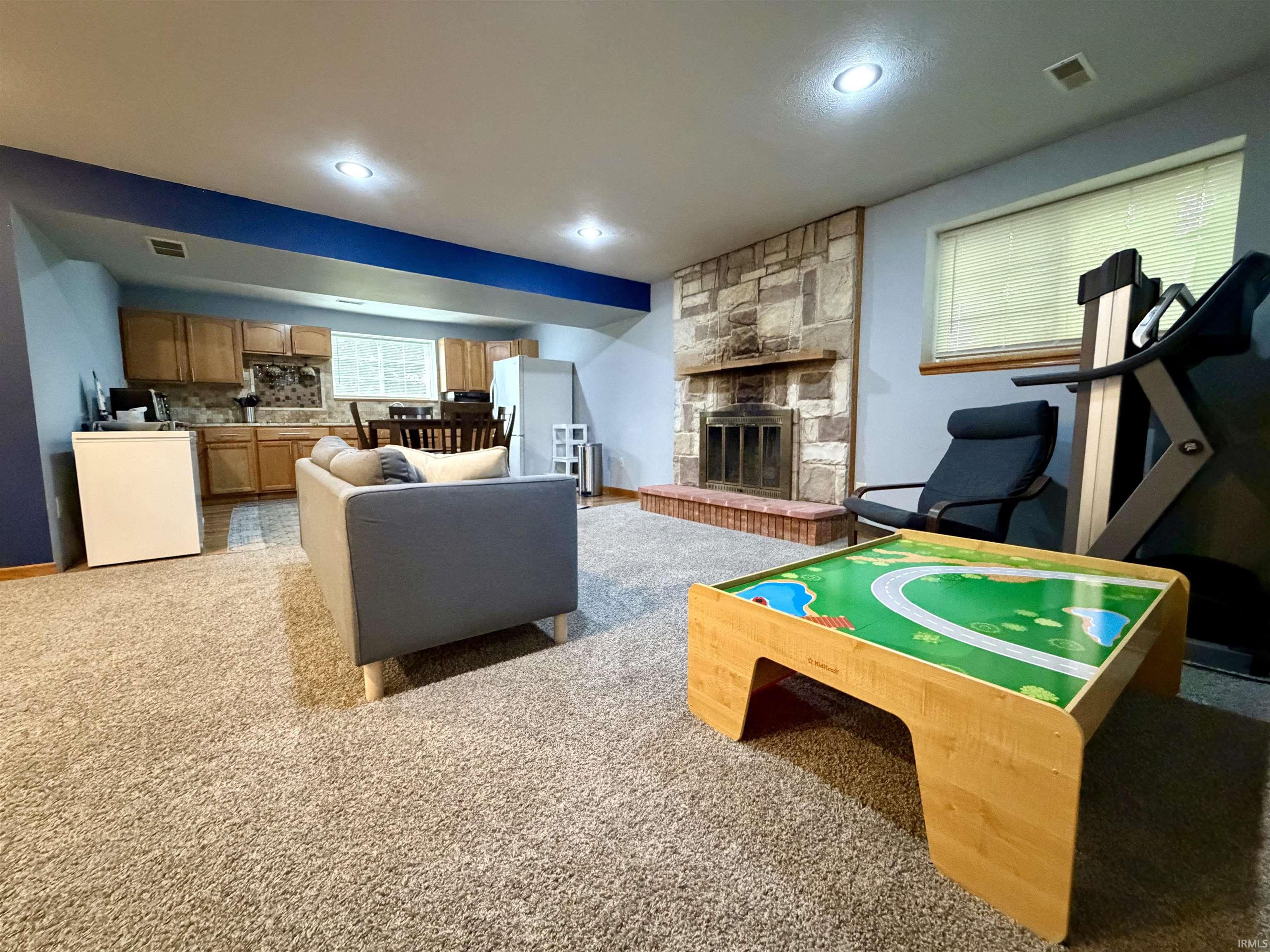 Recessed lighting illuminates a spacious room featuring a stone-clad fireplace with a tiled hearth, a built-in kitchenette with wood cabinetry and a tiled backsplash, and light-toned carpeting