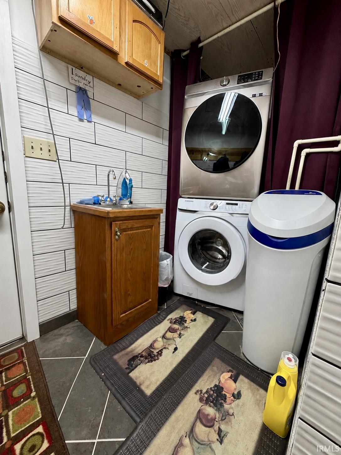 Utility area featuring a laundry sink with wood cabinetry, stacked washer and dryer, tiled walls, and dark flooring
