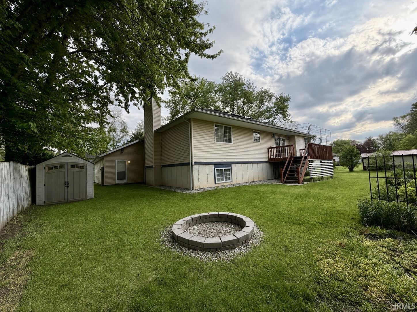 Expansive backyard featuring a stone fire pit, wood deck with stairs, beige vinyl siding, and a detached storage shed