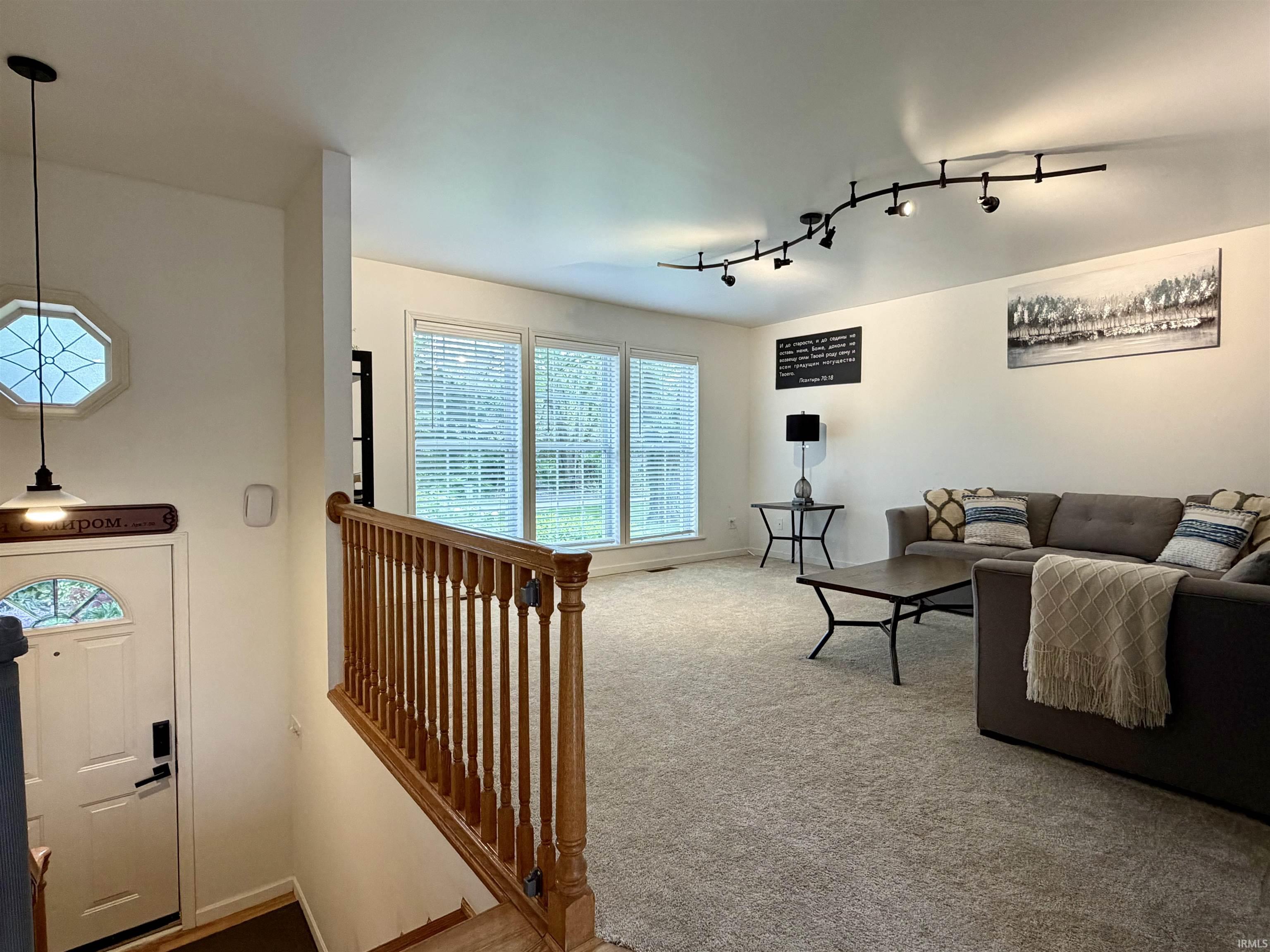 Bright living area featuring track lighting, a wall of three windows with blinds, and light-toned carpet