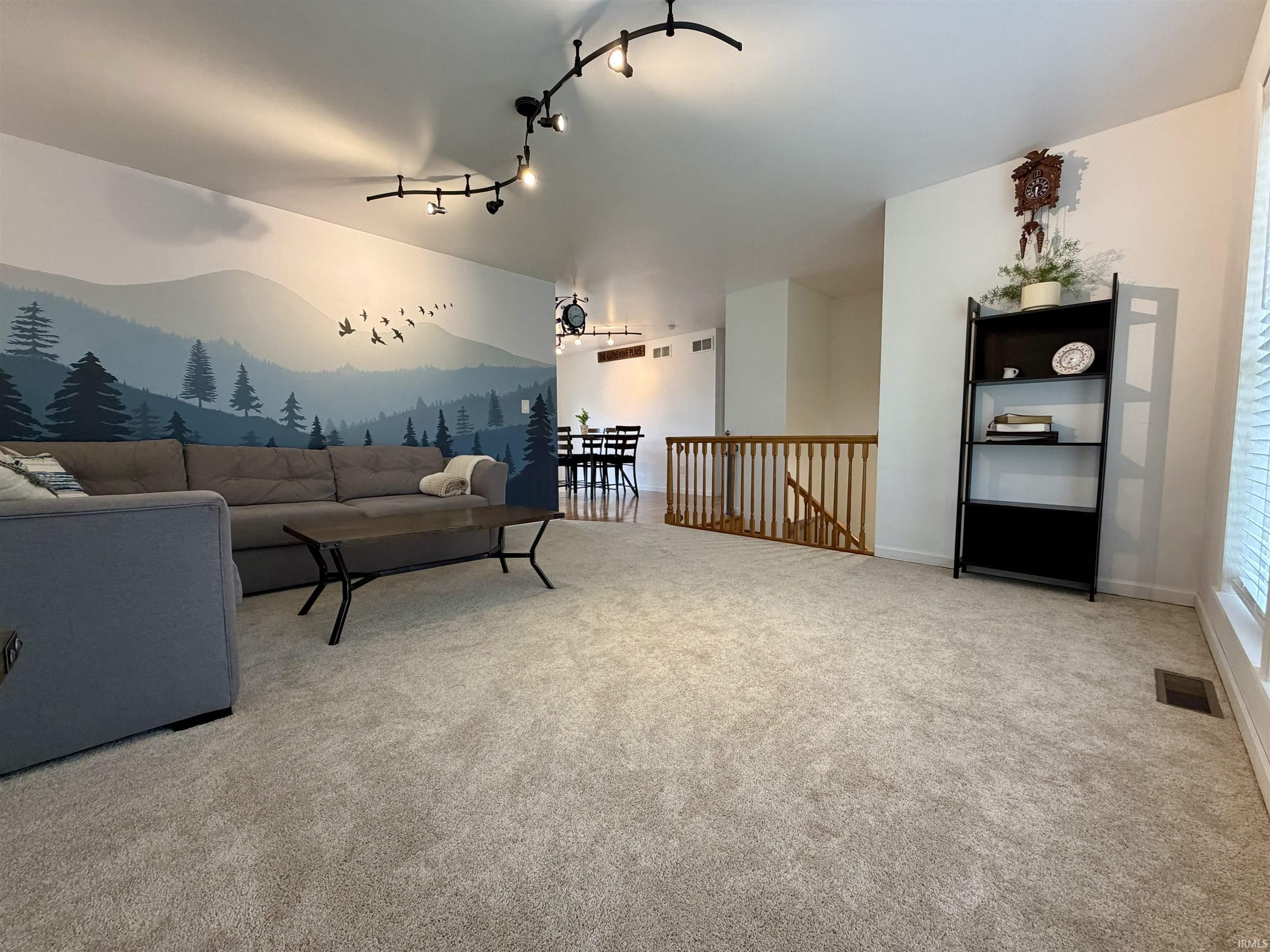 Open-concept living area with light-toned carpeting, track lighting, and a feature wall with a painted mountain mural