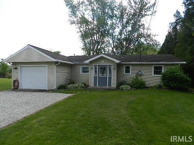 Ranch-style residence featuring light-colored siding, an attached garage, a gravel driveway, a grassy lawn, and mature trees