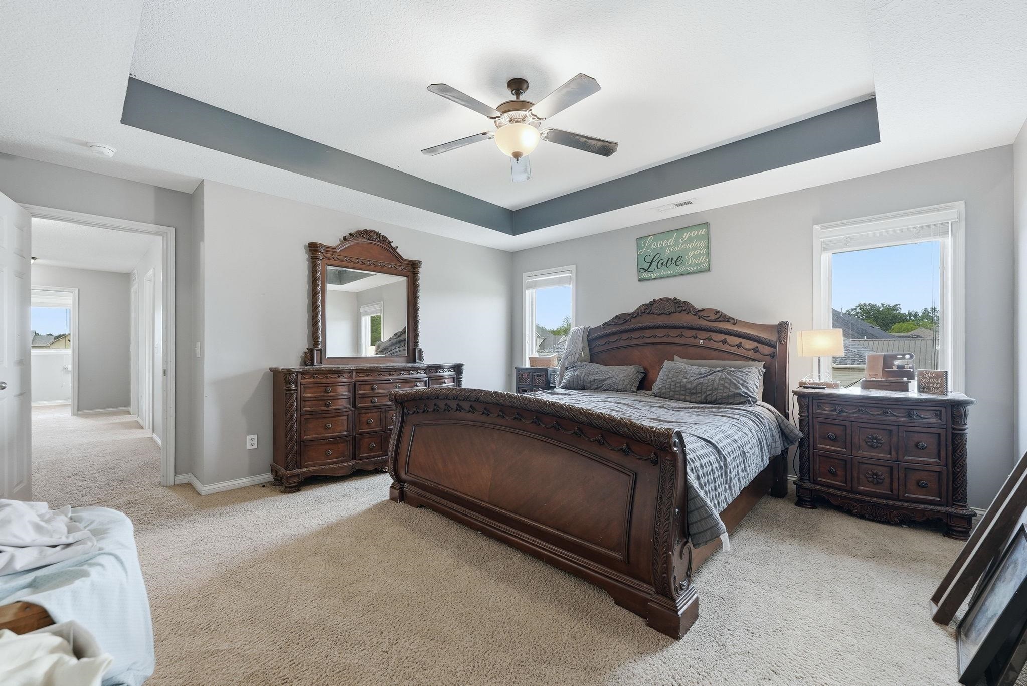 Spacious room featuring a tray ceiling with a ceiling fan, plush carpet flooring, light gray wall paint, and multiple windows for natural illumination