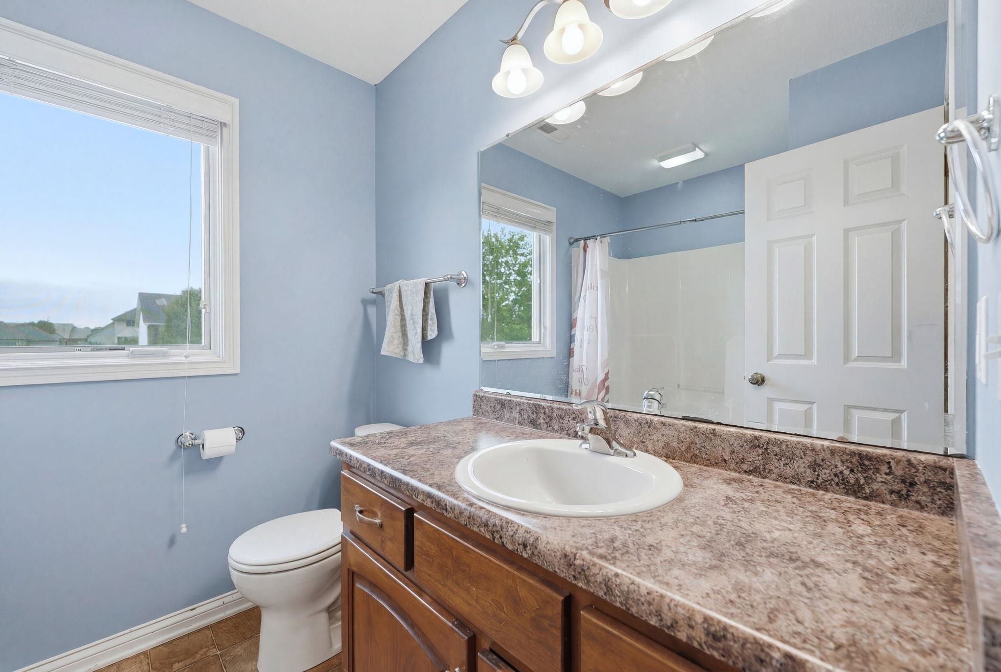 Bathroom featuring a single vanity with a white basin sink, a large mirror, a built-in bathtub with shower, a toilet, and a window with blinds