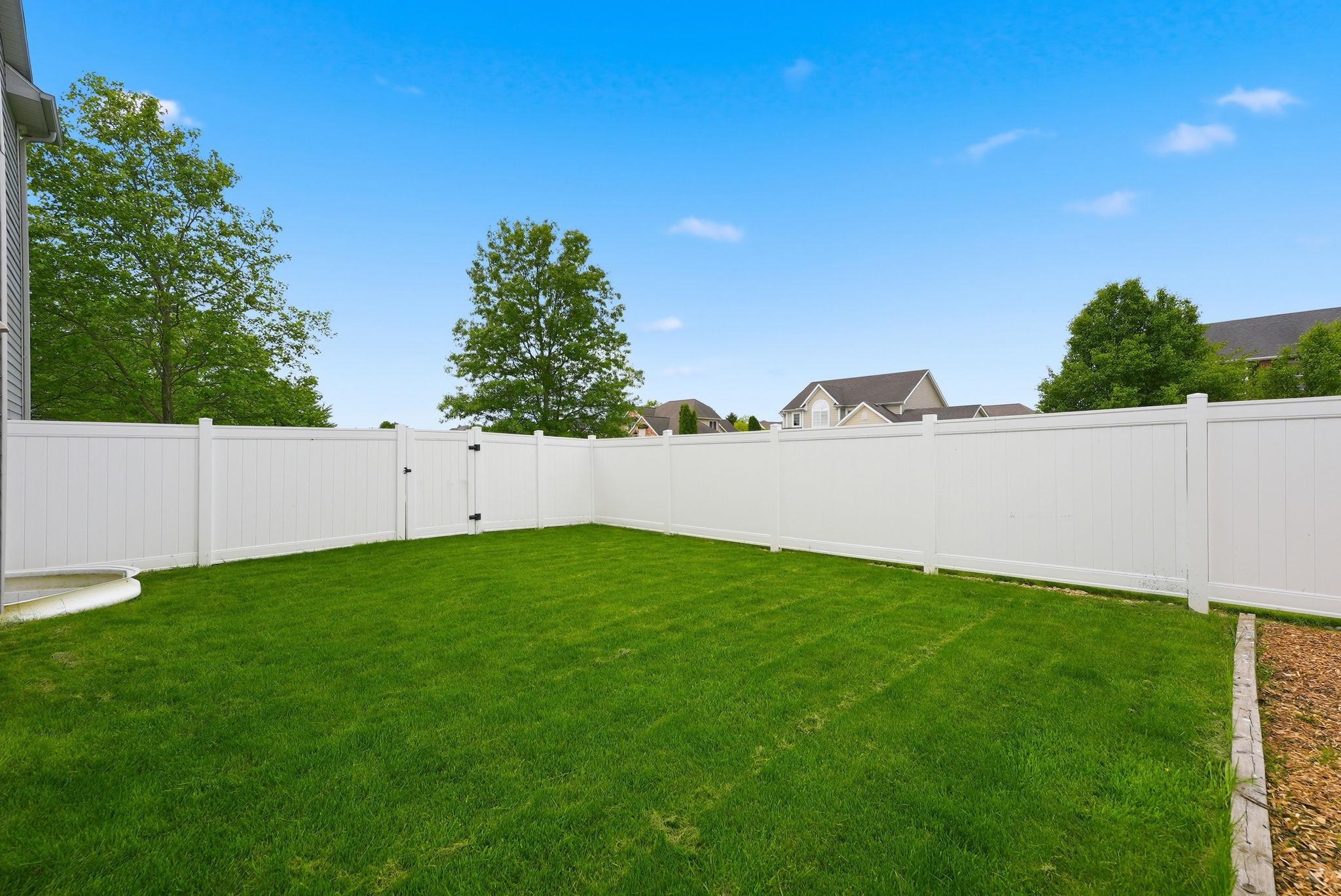 Expansive grassy yard enclosed by a white privacy fence with a gated entry, featuring a curved concrete patio section and an adjacent gravel area