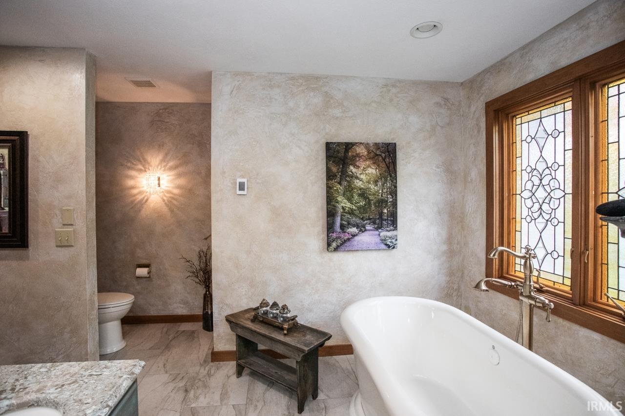 Freestanding soaking tub with floor-mounted faucet hardware, stained-wood framed windows with decorative leaded glass, and light-toned marble-look flooring