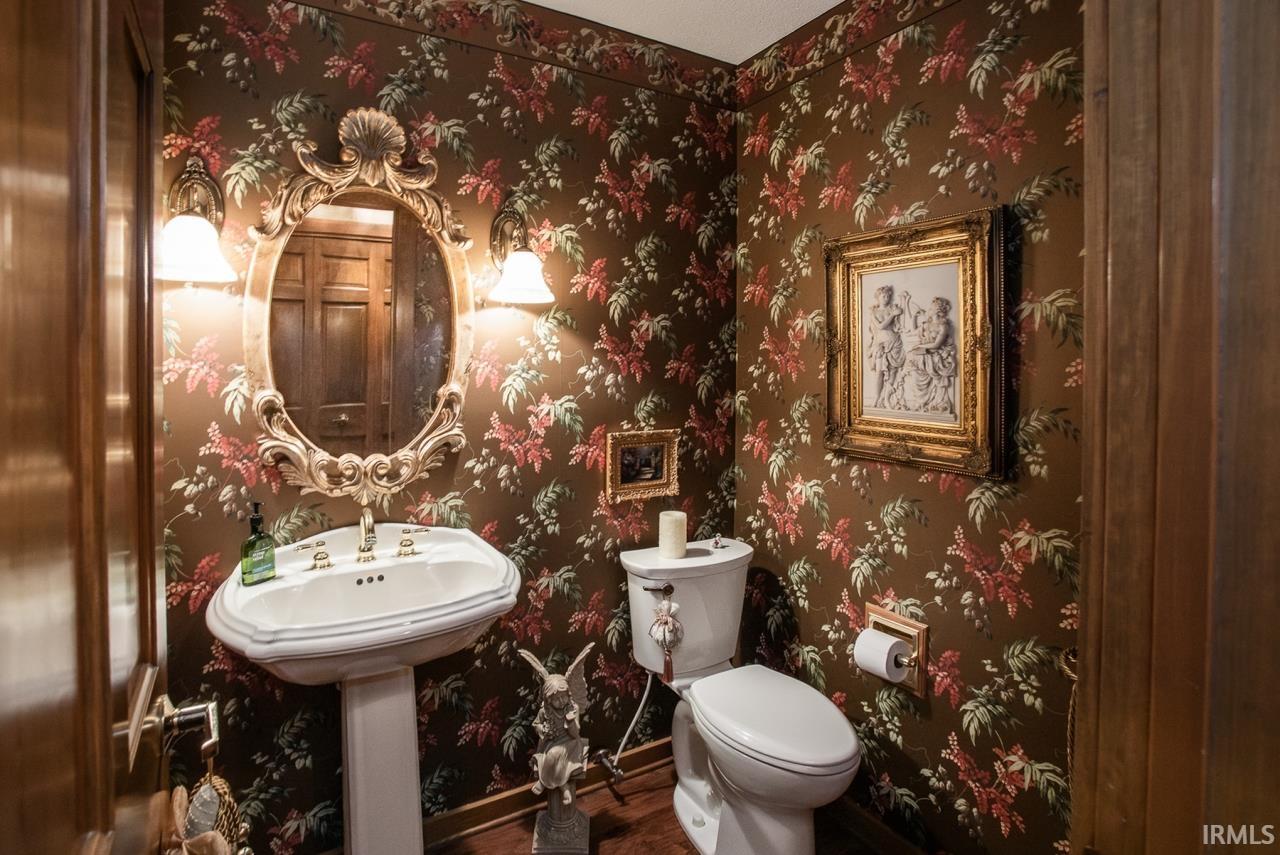 Powder room featuring botanical print wallpaper, a white pedestal sink with brass-toned fixtures, a decorative oval mirror with integrated sconce lighting, and wood-finish flooring