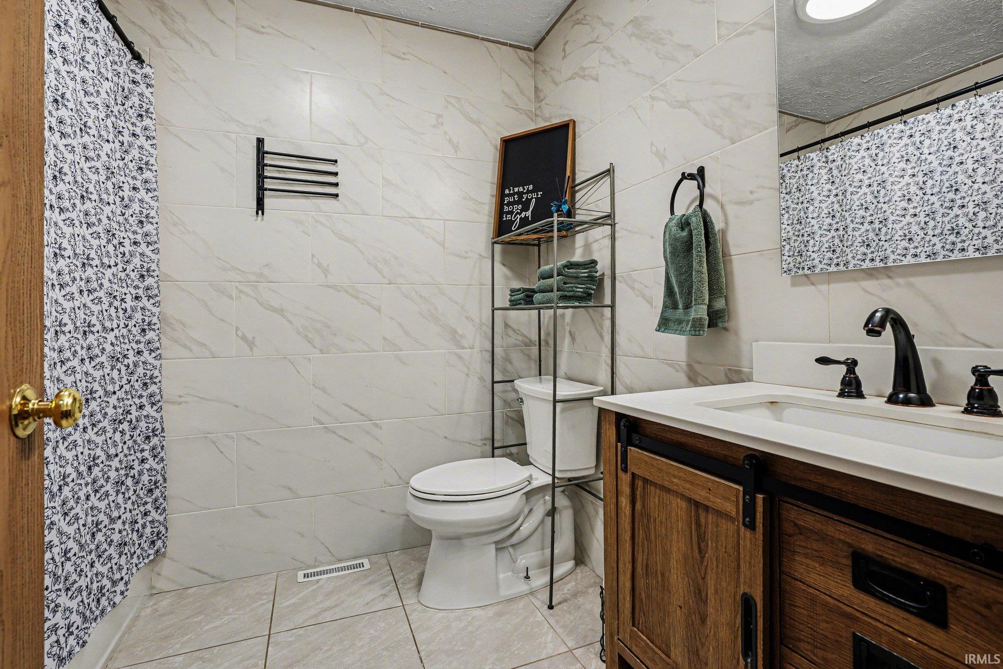 Bathroom featuring white-veined tile walls, a toilet, and a wood-finish vanity with a white countertop and matte black fixtures