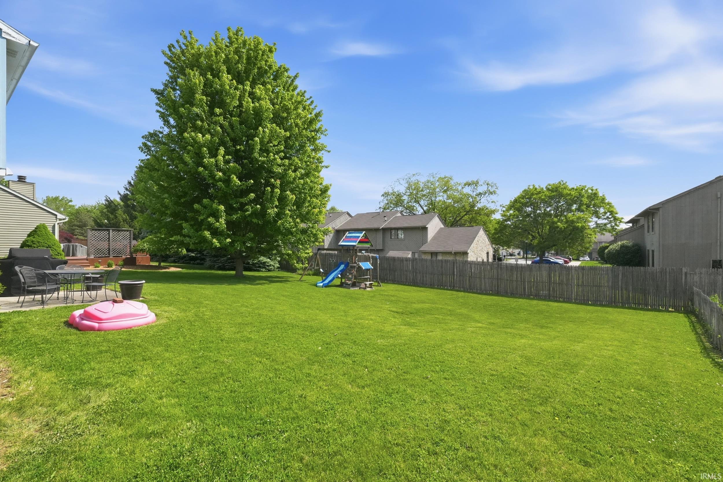 Expansive rear yard featuring a large grass lawn, mature trees, a wooden privacy fence, a concrete patio, and a raised wooden deck