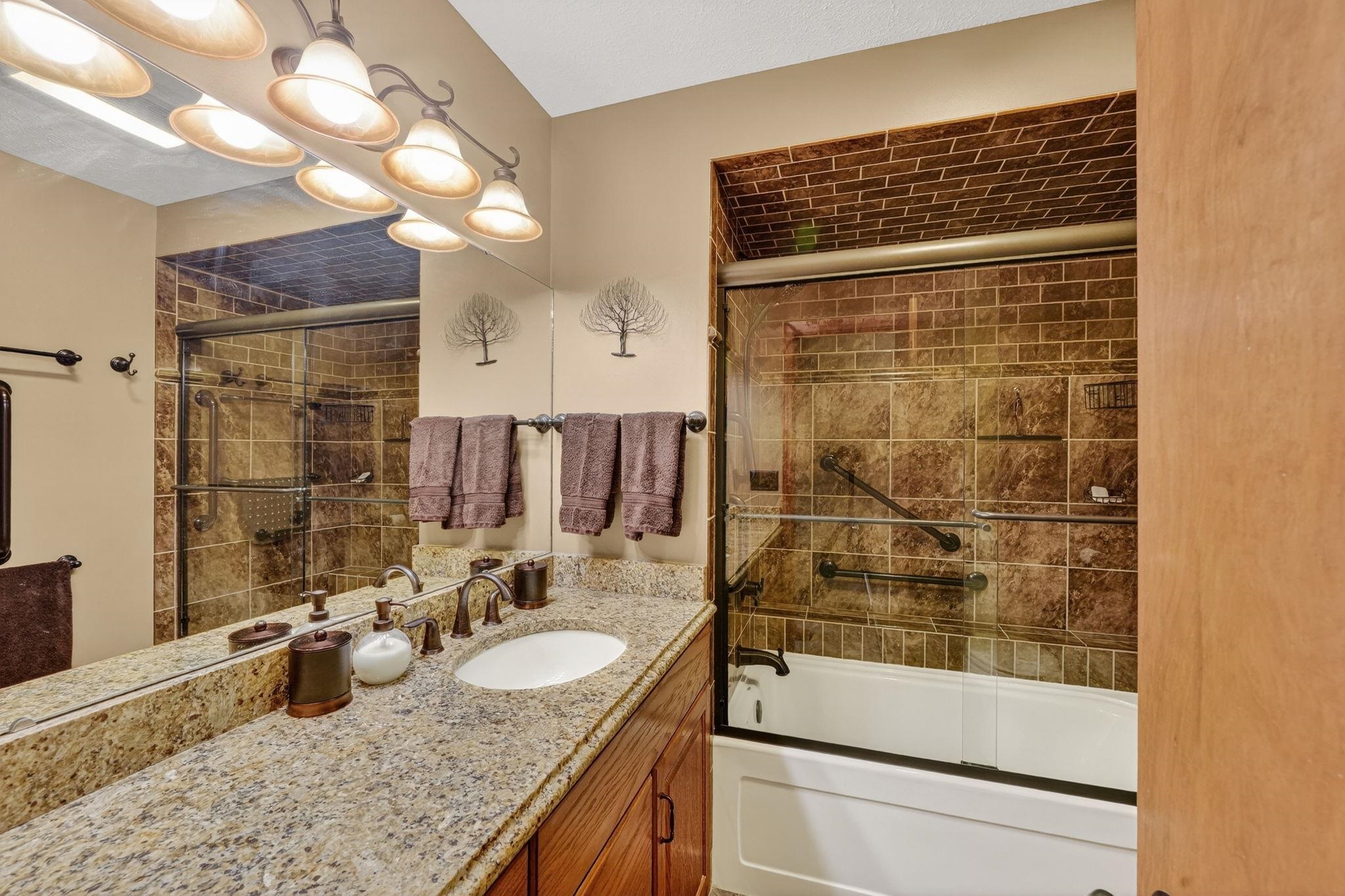 Bathroom featuring a granite-finish countertop, wood-finish cabinetry, a large vanity mirror, and a full-size bathtub with tiled surround and glass sliding doors