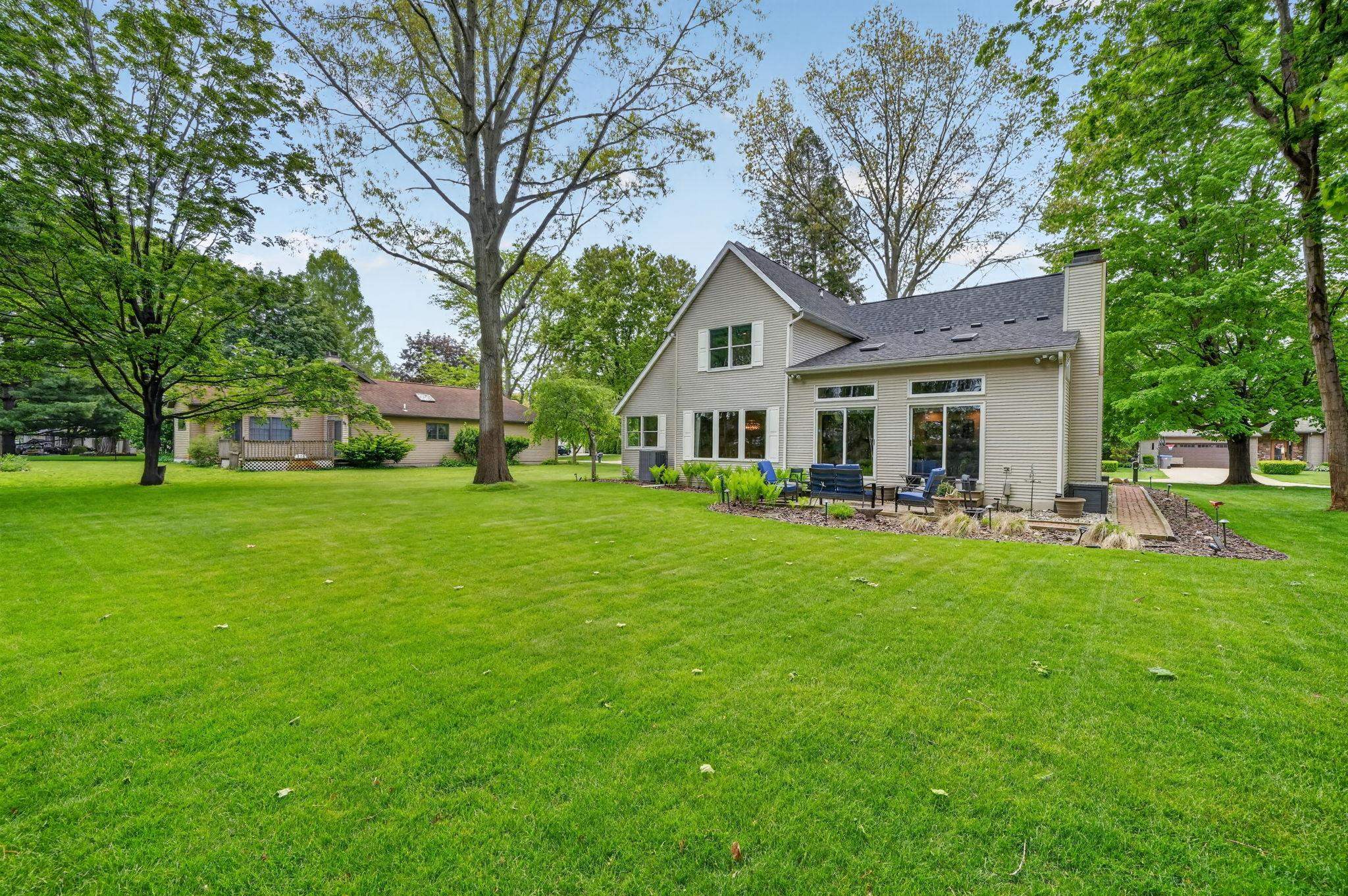 Expansive green lawn surrounds a property featuring multiple large windows and glass sliding doors, a grey shingle roof, and a stone chimney