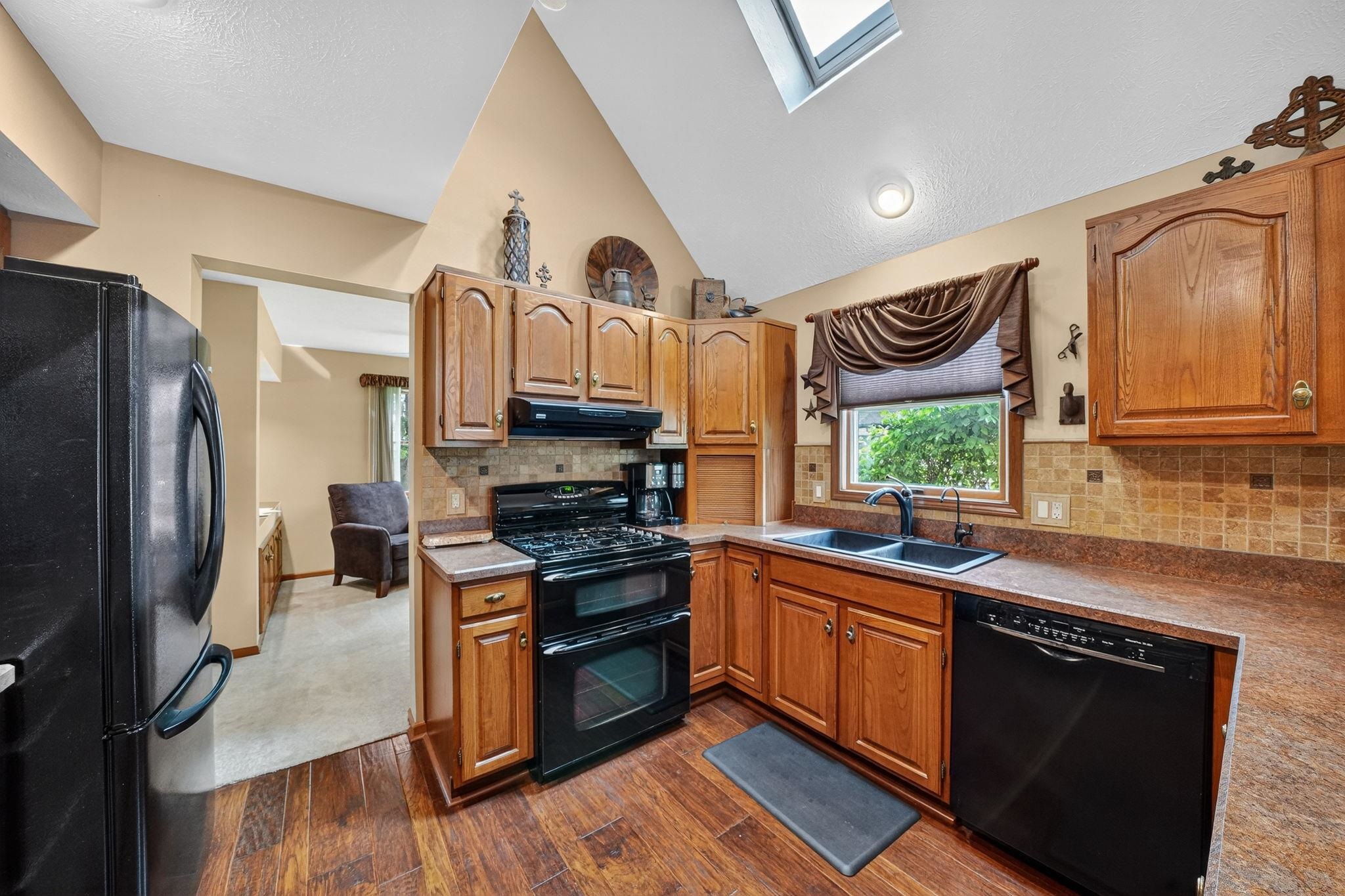 Kitchen featuring wood-finish flooring, a double-basin sink, black appliances, a tile backsplash, and a skylight