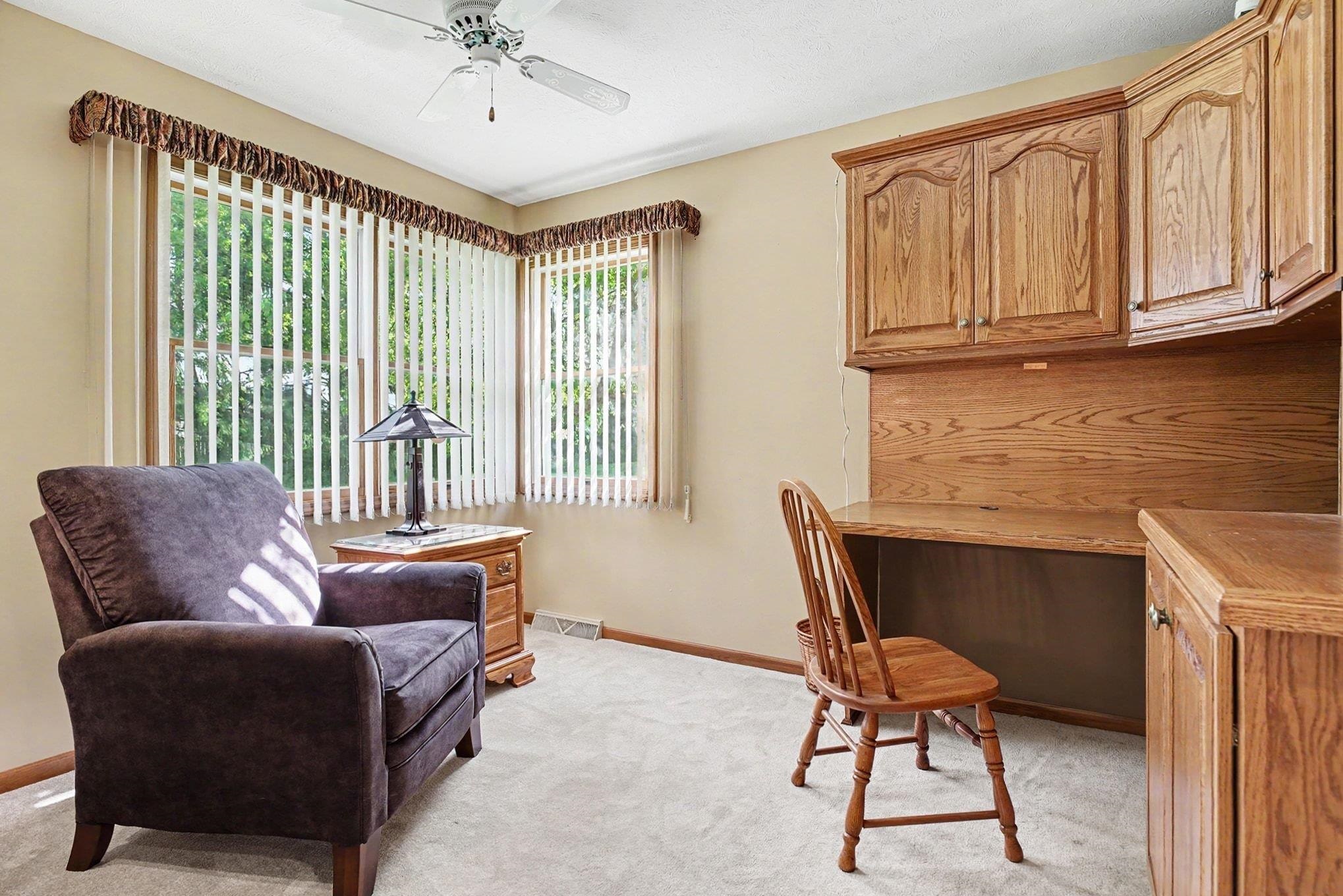 Room featuring a built-in wood-finish desk with overhead cabinetry, multiple windows with vertical blinds, and a ceiling fan